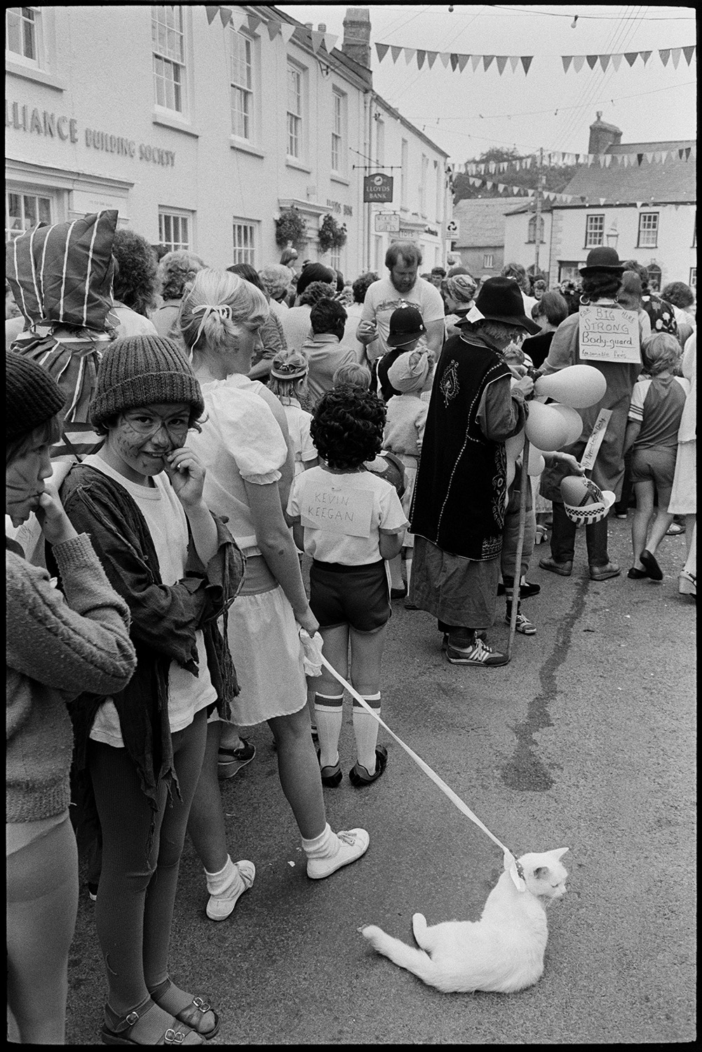 Fancy dress parade with brass band, going home afterwards. 
[A young woman with a cat on a lead at Chulmleigh Fair. Other people can be seen, including children with their faces painted, people in fancy dress and a person with balloons. The street is decorated with bunting.]