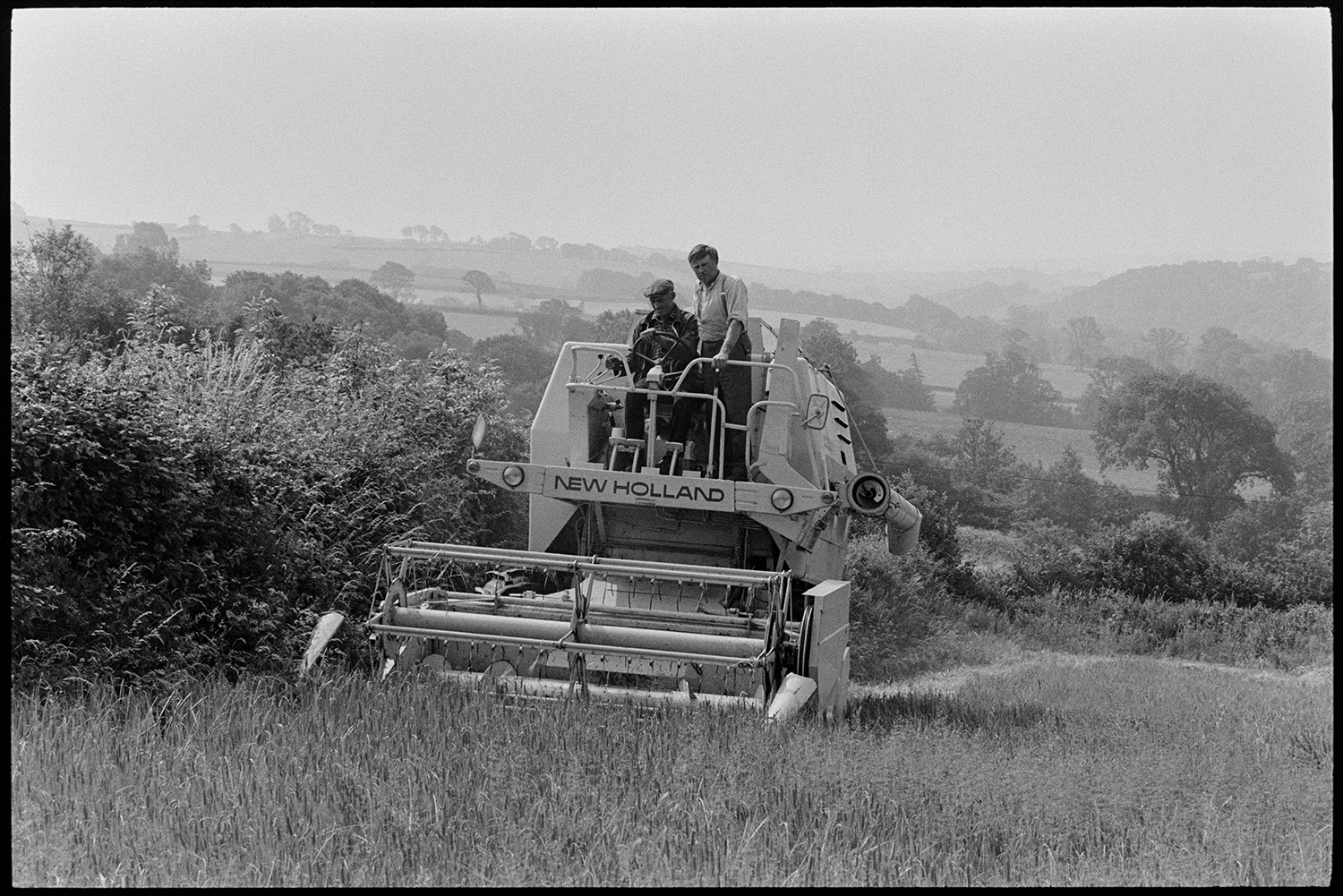 Combine harvester working in landscape. 
[Two men driving a combine harvester alongside a hedge in a field at Brightley, Dolton, to harvest a crop. A landscape of trees and fields can be seen in the background.]