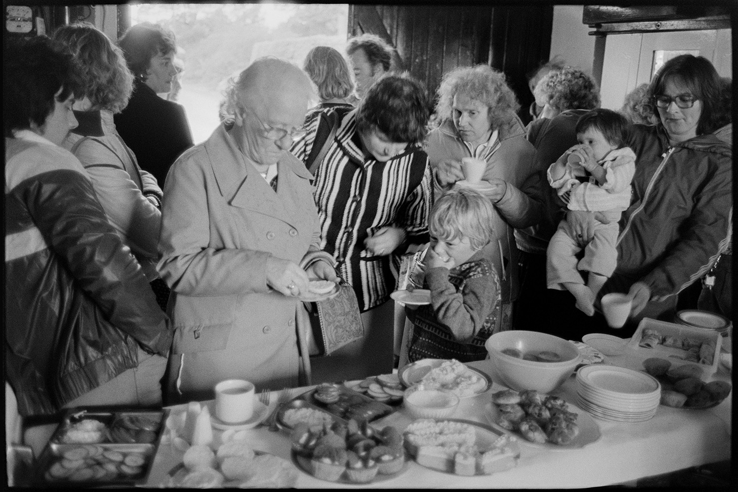 Vicarage fete, stalls, croquet, tea and cakes in rain in garden, dogs. 
[Men, women and children helping themselves to tea, cakes and biscuits in a garage, at the vicarage fete at Merton Rectory.]