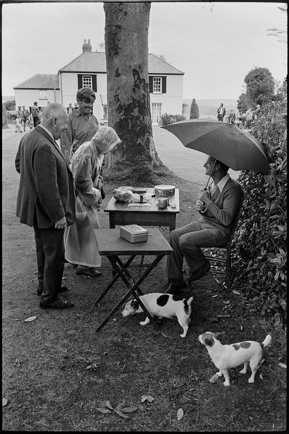 Vicarage fete, stalls, croquet, tea and cakes in rain in garden, dogs. 
[People arriving and having a go on the raffle at the vicarage fete at Merton Rectory. A man with an umbrella and two dogs are at the entrance. The vicarage can be seen in the background.]