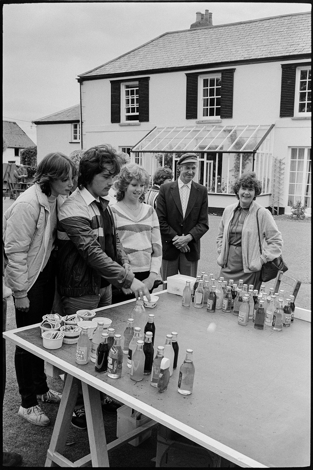 Vicarage fete, stalls, croquet, tea and cakes in rain in garden, dogs. 
[A man trying to win a prize bottle by throwing a ping pong ball into a jar at the vicarage fete at Merton Rectory. Other people are watching him. The vicarage can be seen in the background.]
