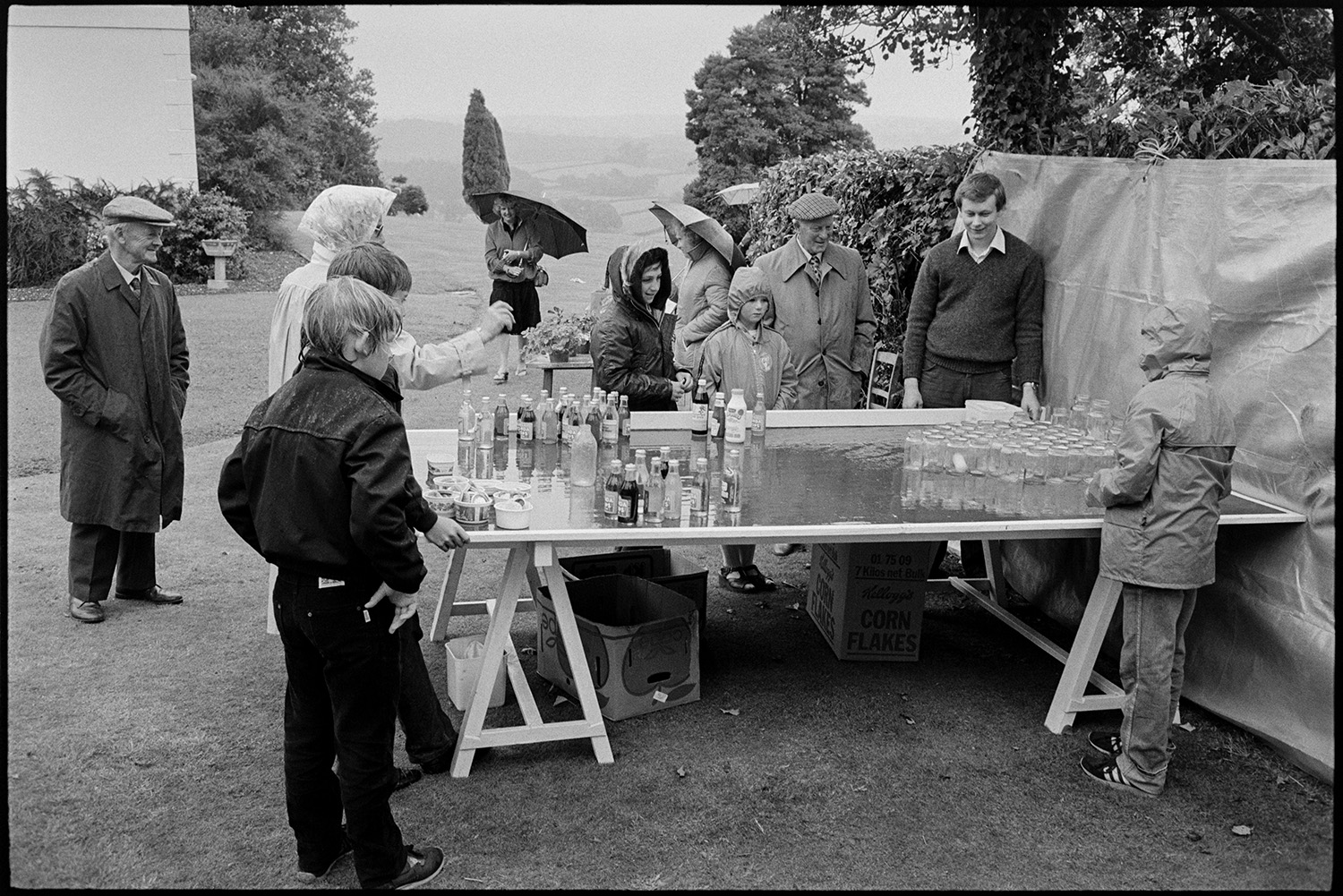 Vicarage fete, throwing the ping pong ball into the jar. Umbrellas in rain in garden. 
[A woman trying to win a prize bottle by throwing a ping pong ball in a jar, in the rain at the vicarage fete at Merton Rectory. Children and men are watching her. Two women are holding umbrellas in the background.]