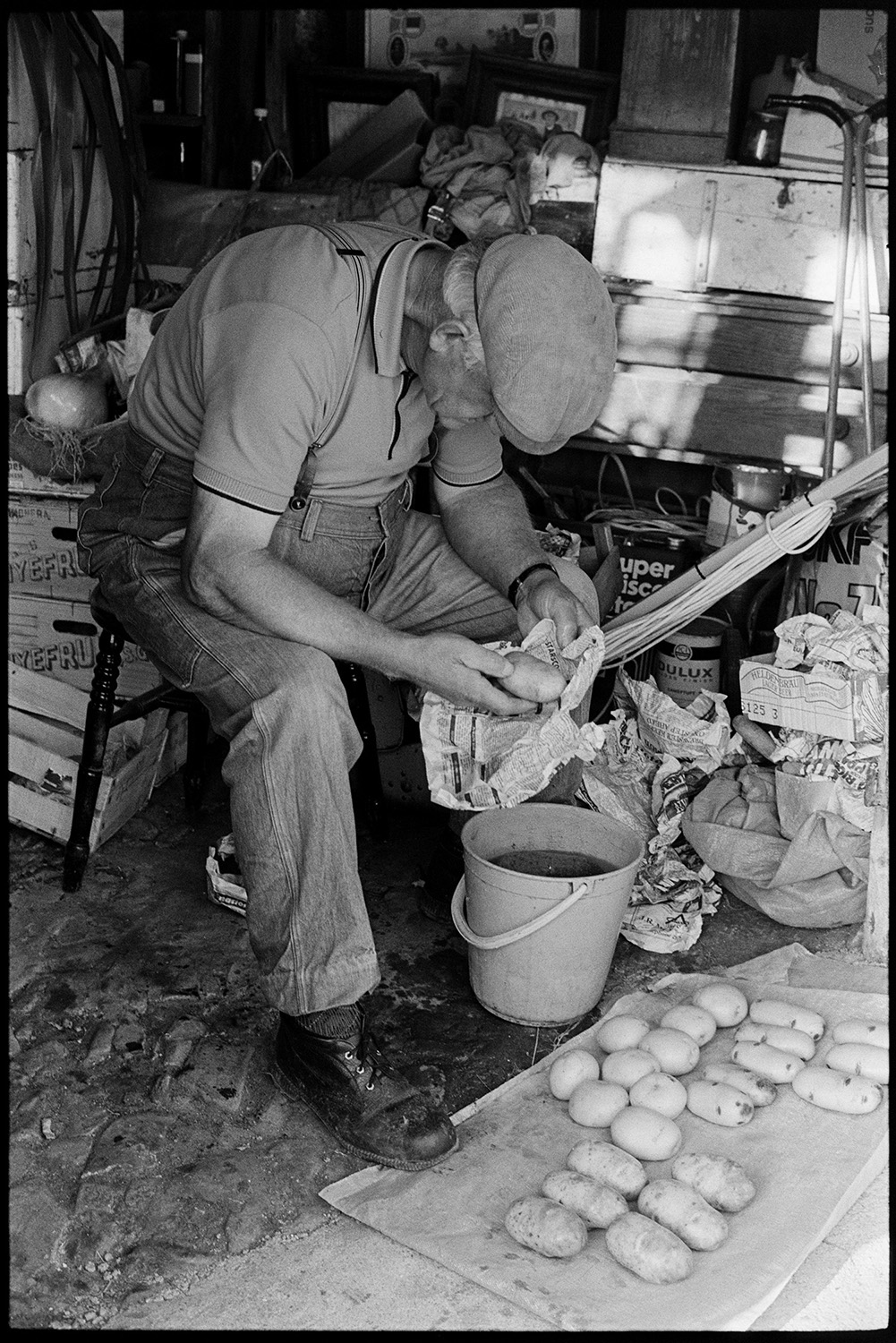 Father and son with vegetables for flower show, digging up parsnips, leeks, onions in shed. 
[Lloyd Mitchell washing potatoes in a bucket in a shed with a cobbled floor at Pear Tree Cottage, Aller Road, Dolton. He is preparing the vegetables for Dolton Flower Show.]