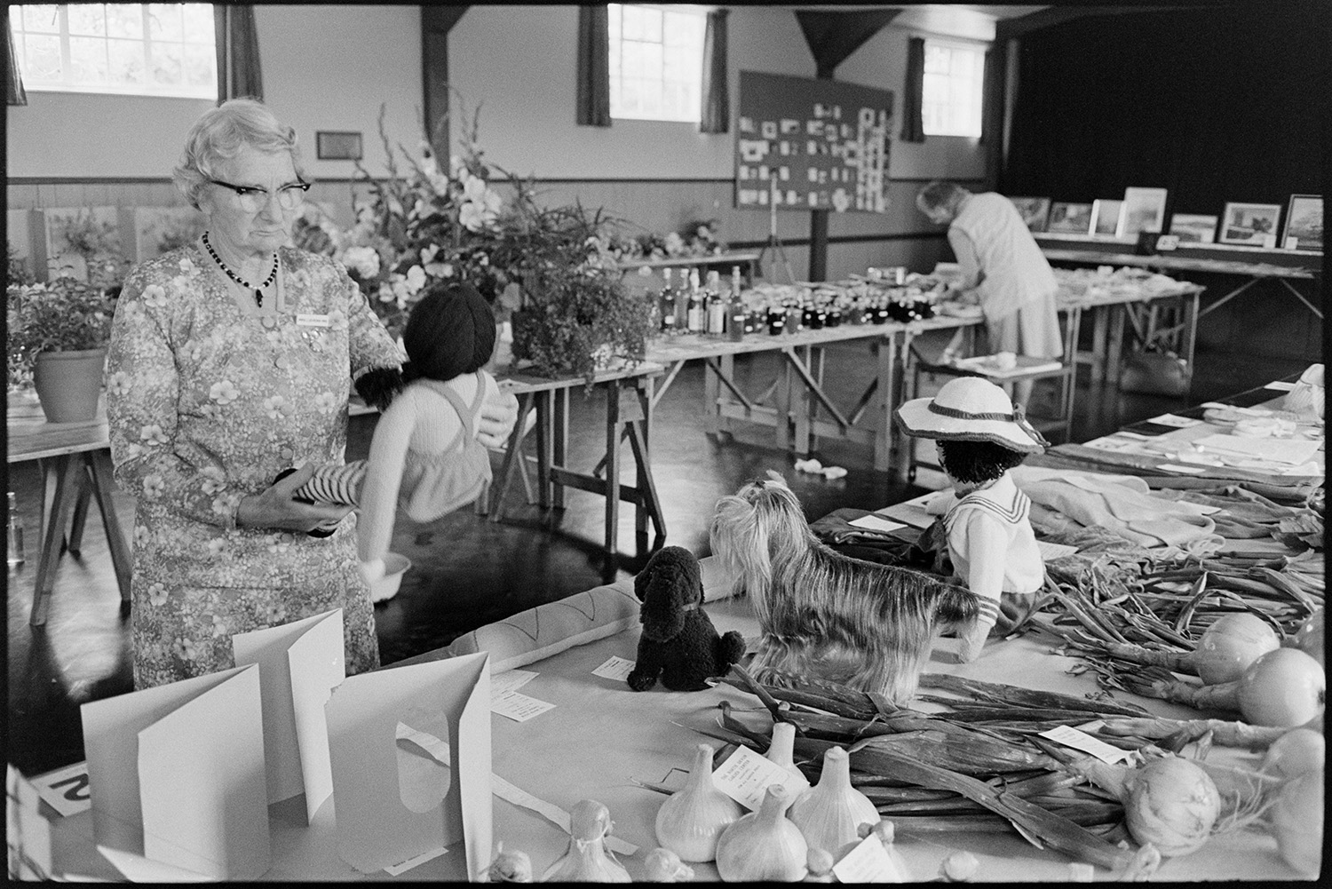 Vegetables, produce and cakes at flower show in village hall, French beans, onions, flowers.
[Woman judging exhibits at Dolton Flower Show in Dolton Village Hall, including a knitted doll. There are tables of flowers, vegetables, bottles and jams on display.]
