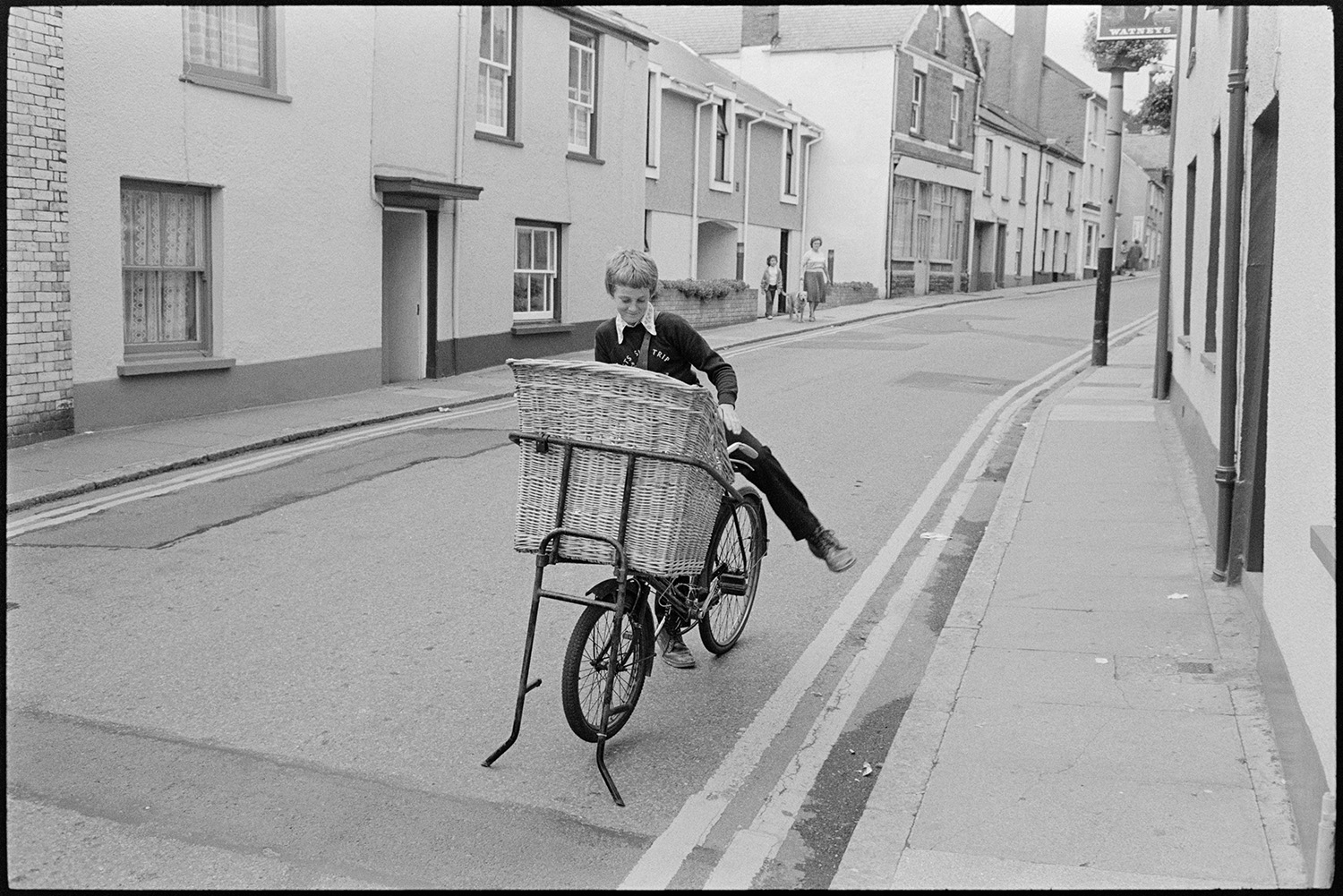 Delivery boy on bicycle with basket in street.
[A delivery boy on a bicycle with a large basket, in Well Street, Torrington.]