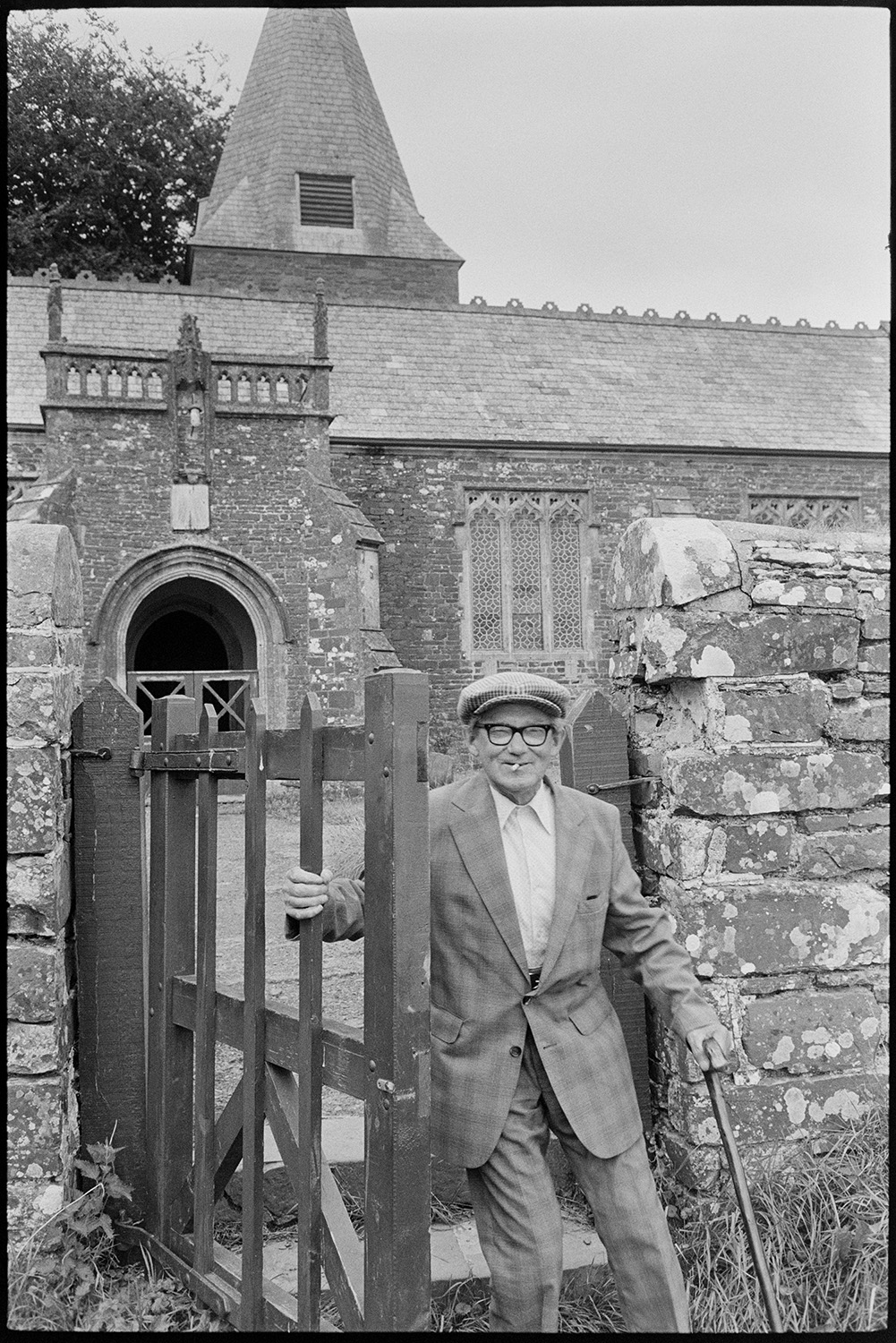 Village revel, fete, women at cake stall, guessing weight of ram, side shows.
[A man walking through the gate from Beaford churchyard, wearing a suit and smoking a cigarette. He is using a walking stick. Beaford Church can be seen in the background.]
