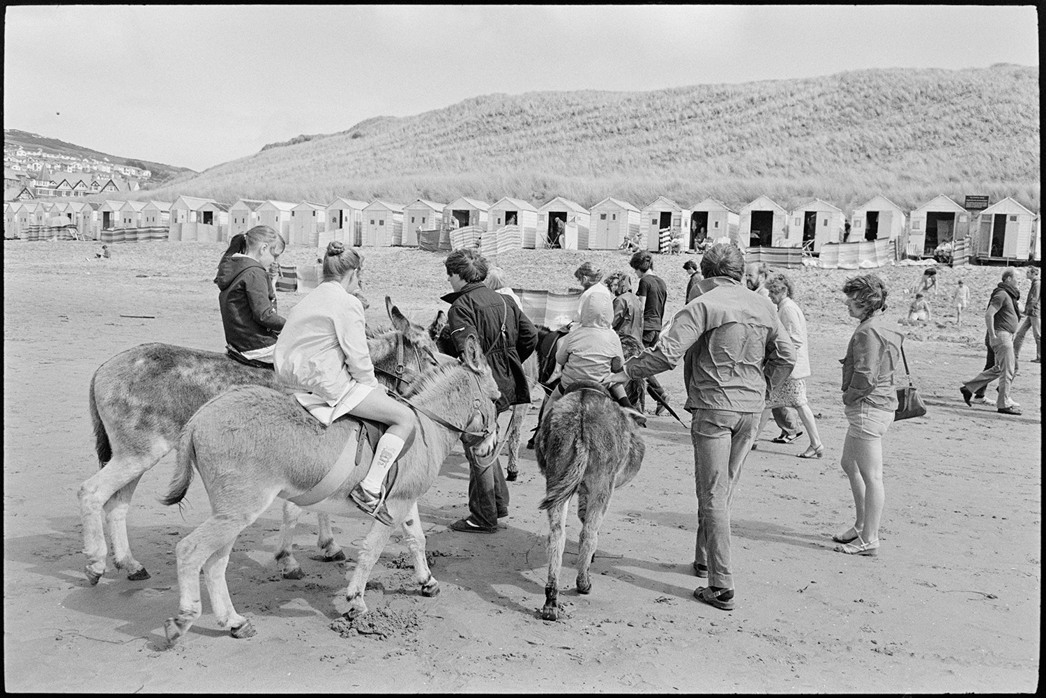 Beach scenes with donkey rides, bathing huts, people swimming.
[Children having donkey rides on Woolacombe beach. Sand dunes, holidaymakers and beach huts are visible in the background.]