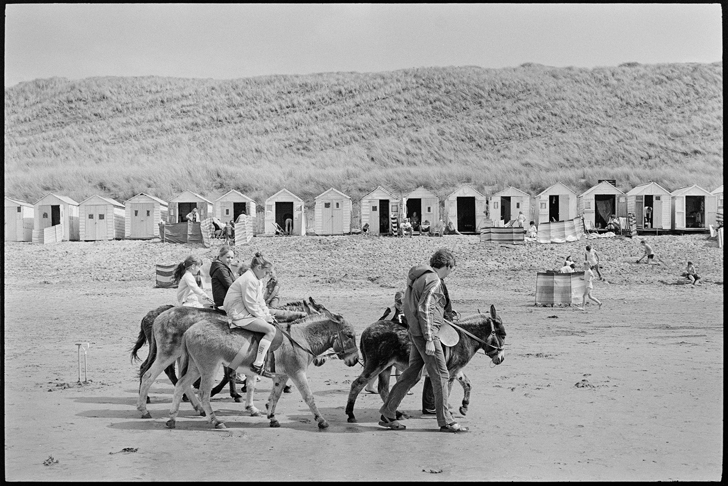 Beach scenes with donkey rides, bathing huts, people swimming.
[Children having donkey rides on Woolacombe beach. Sand dunes, holidaymakers sat behind wind breaks and beach huts are visible in the background.]