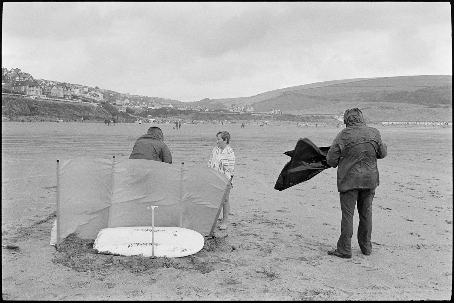 Beach scenes with donkey rides, bathing huts, people swimming.
[Holidaymakers on a wet and windy Woolacombe beach.  A surfboard is resting behind a windbreak and a child is wrapped in a towel. The town of Woolacombe is visible in the background.]