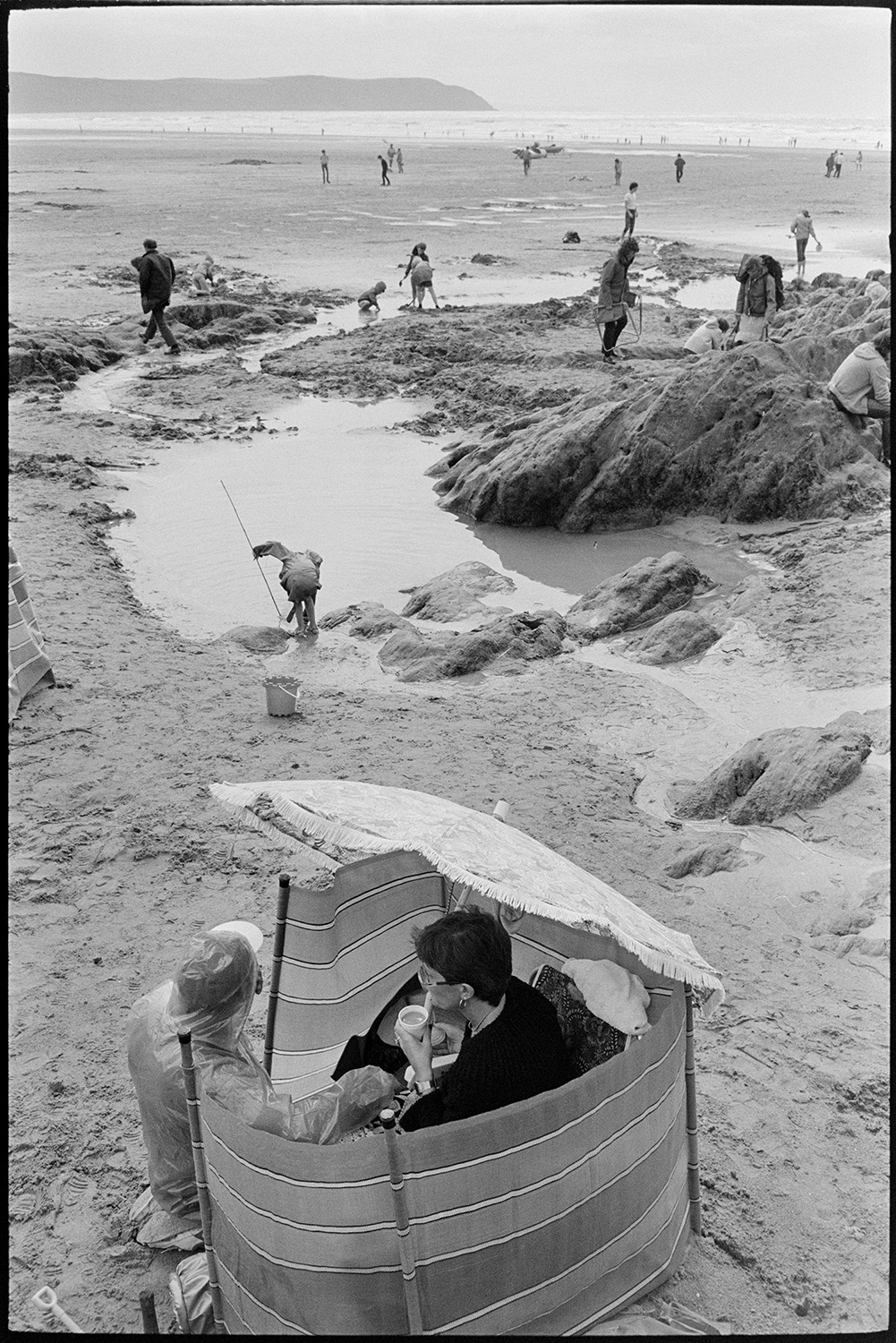 Beach scenes with people sheltering and having tea, children playing in rock pools.
[Holidaymakers on Woolacombe beach sheltering behind a windbreak and drinking tea. Children are playing in rock pools nearby and the sea and cliffs are visible in the background.]