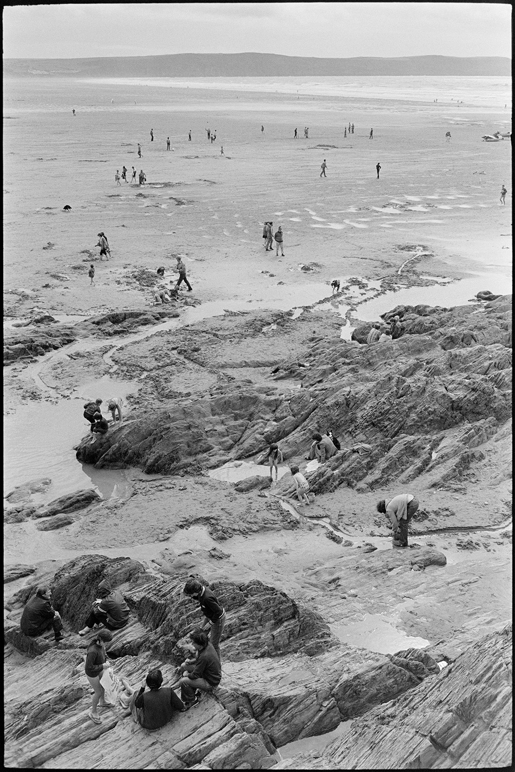 Beach scenes with people sheltering and having tea, children playing in rock pools.
[Holidaymakers sitting on rocks at Woolacombe beach, with children playing in rock pools nearby. The sea and people walking on the beach are visible in the background.]