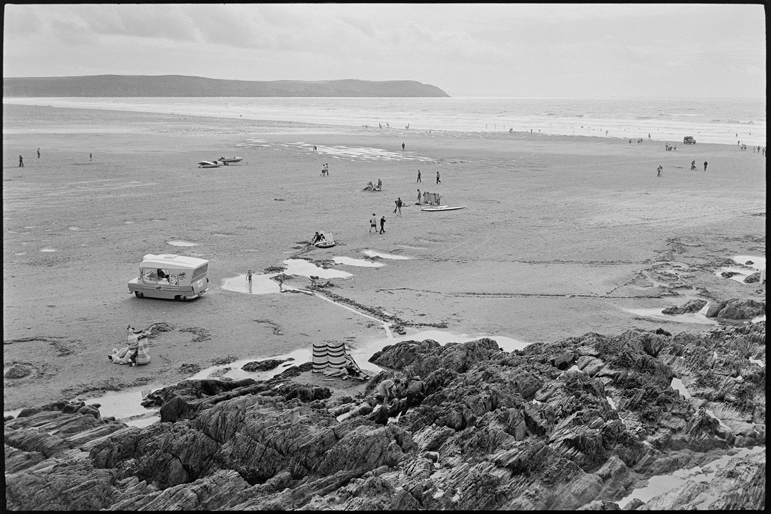 Beach scenes with people sheltering and having tea, children playing in rock pools.
[Holidaymakers and an ice cream van on Woolacombe beach. Some people are sat behind windbreaks by rocks in the foreground. Other people are in the sea in the background.]