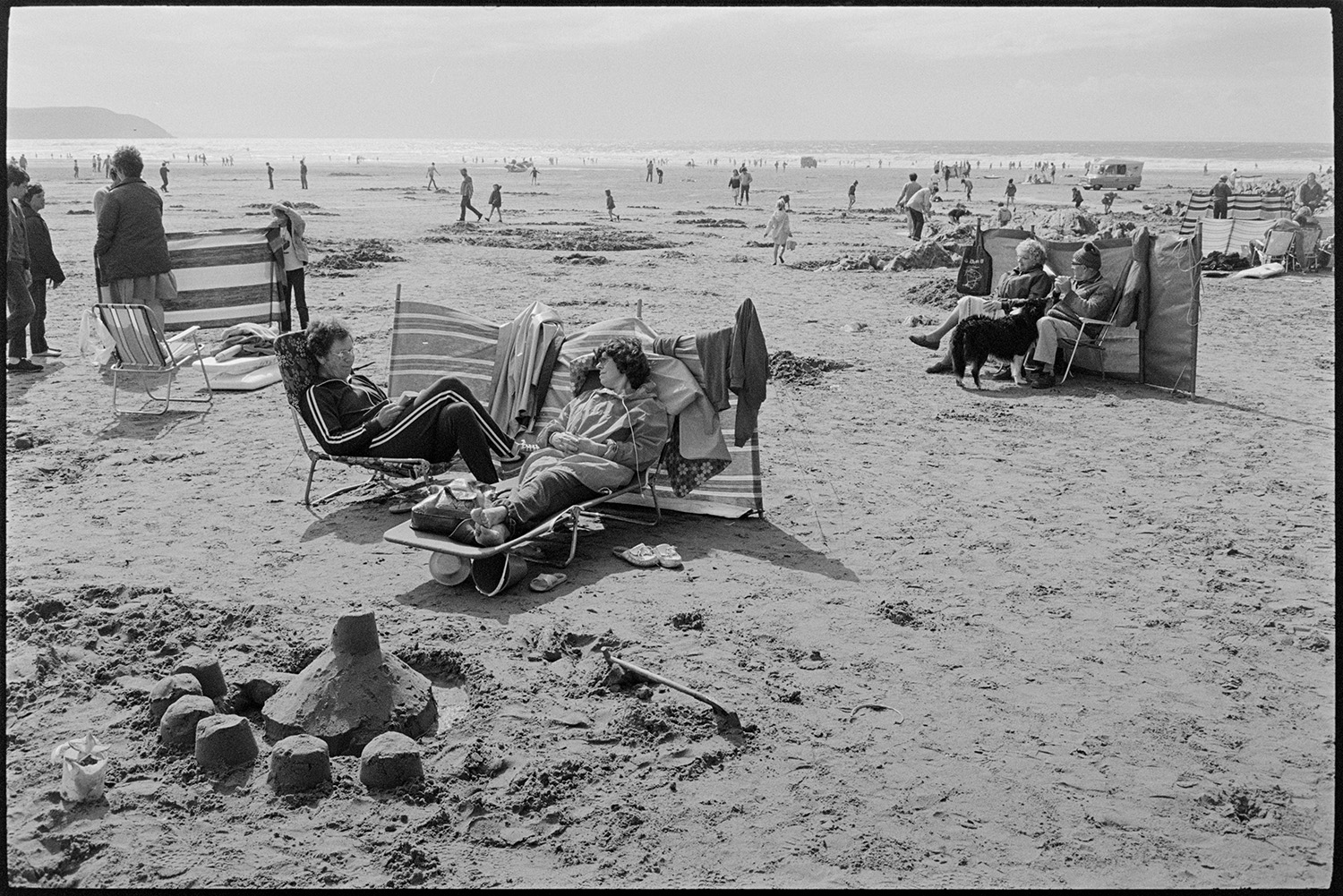 Beach scenes, people sitting behind windbreaks.
[Holidaymakers on Woolacombe beach sitting behind windbreaks, with sand castles in the foreground. An ice cream van and other holidaymakers are visible in the distance.]