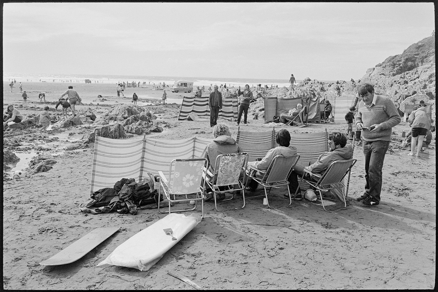 Beach scenes, people sitting behind windbreaks.
[Holidaymakers on Woolacombe beach sitting on garden chairs behind a windbreak. Surfboards are lying on the beach behind them. Other holidaymakers, an ice cream van and the sea are visible in the background.]
