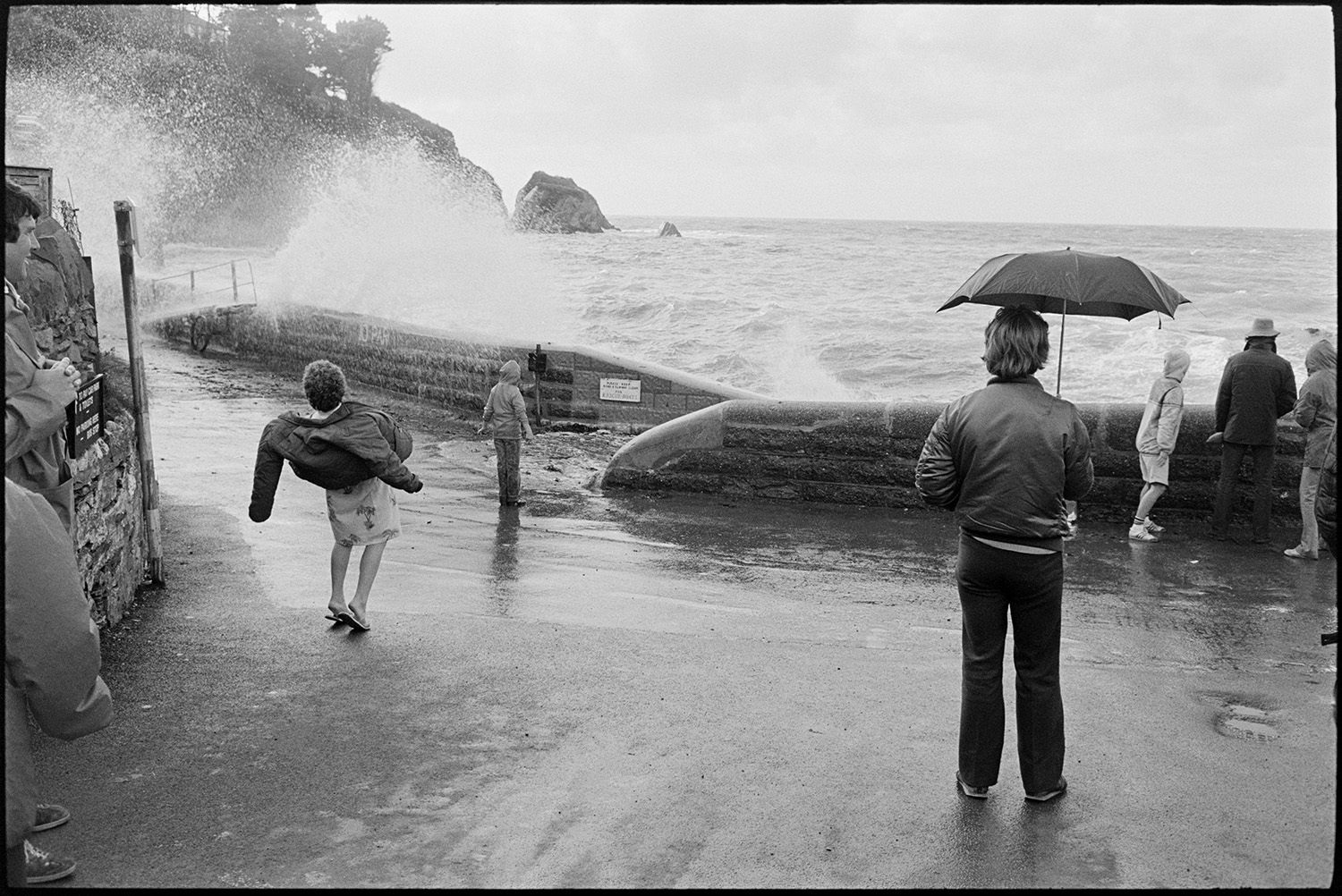 Holidaymakers watching rough seas breaking over sea wall.
[Holidaymakers watching rough seas breaking over a sea wall at Lee Bay. A man is standing holding an umbrella in the foreground and looking at the waves.]
