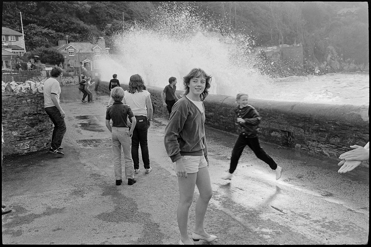 Holidaymakers watching rough seas breaking over sea wall.
[Teenagers watching high waves breaking over the sea wall at Lee Bay. Houses are visible on the seafront in the background.]