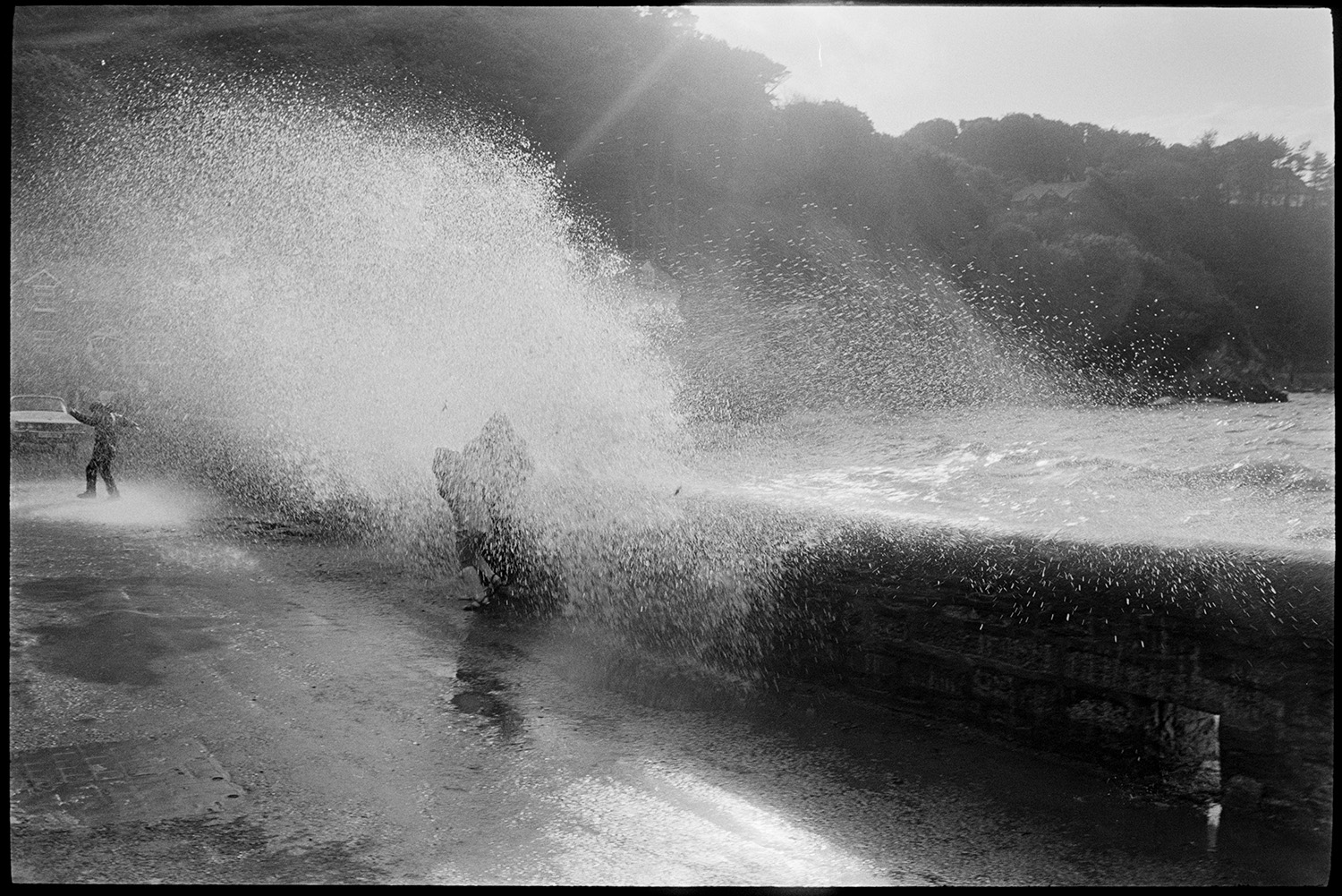 Holidaymakers watching rough seas breaking over sea wall.
[A wave breaking over the sea wall at Lee Bay. People and a car are being splashed by the wave. Wooded cliffs are visible in the background.]