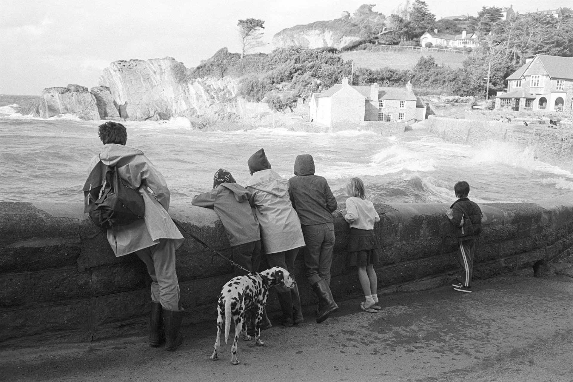 Holidaymakers watching rough seas across bay, leaning on sea wall, dog. 
[Holidaymakers leaning on the sea wall at Lee Bay and watching waves crashing. Cliffs and buildings can be seen across the bay. One of the holidaymakers has a dog.]