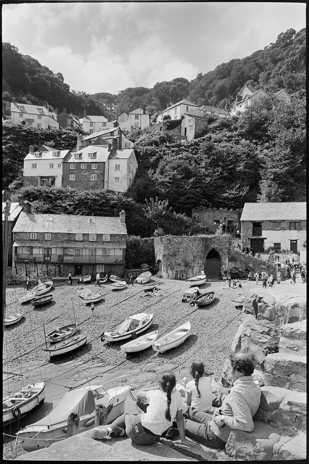 Holidaymakers, seaside village taking photographs, sunbathing, pebble beach with small boats. 
[Holidaymakers sitting on the harbour wall overlooking the beach at Clovelly. Lots of small boats are moored on the pebble beach and houses can be seen on the valley hillside in the background.]