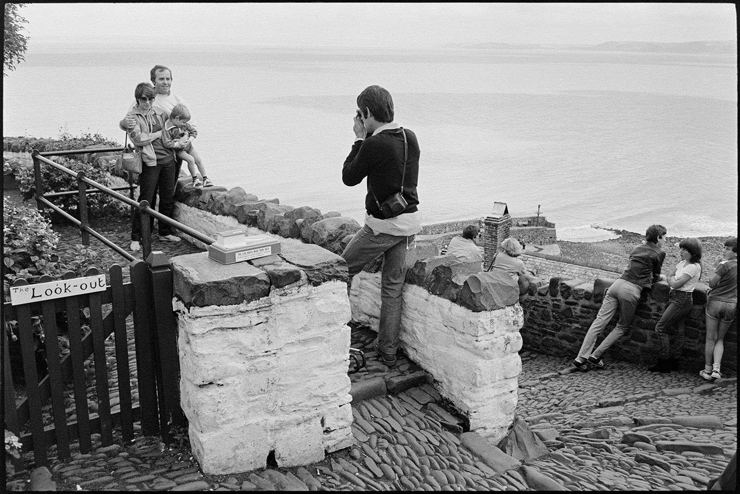 Holidaymakers, seaside village taking photographs, sunbathing, pebble beach with small boats. 
[A person taking a photograph of a family at a look out point at Clovelly. The cobbled steps leading down the to beach are visible, as well as part of the harbour wall and sea, in the background.]