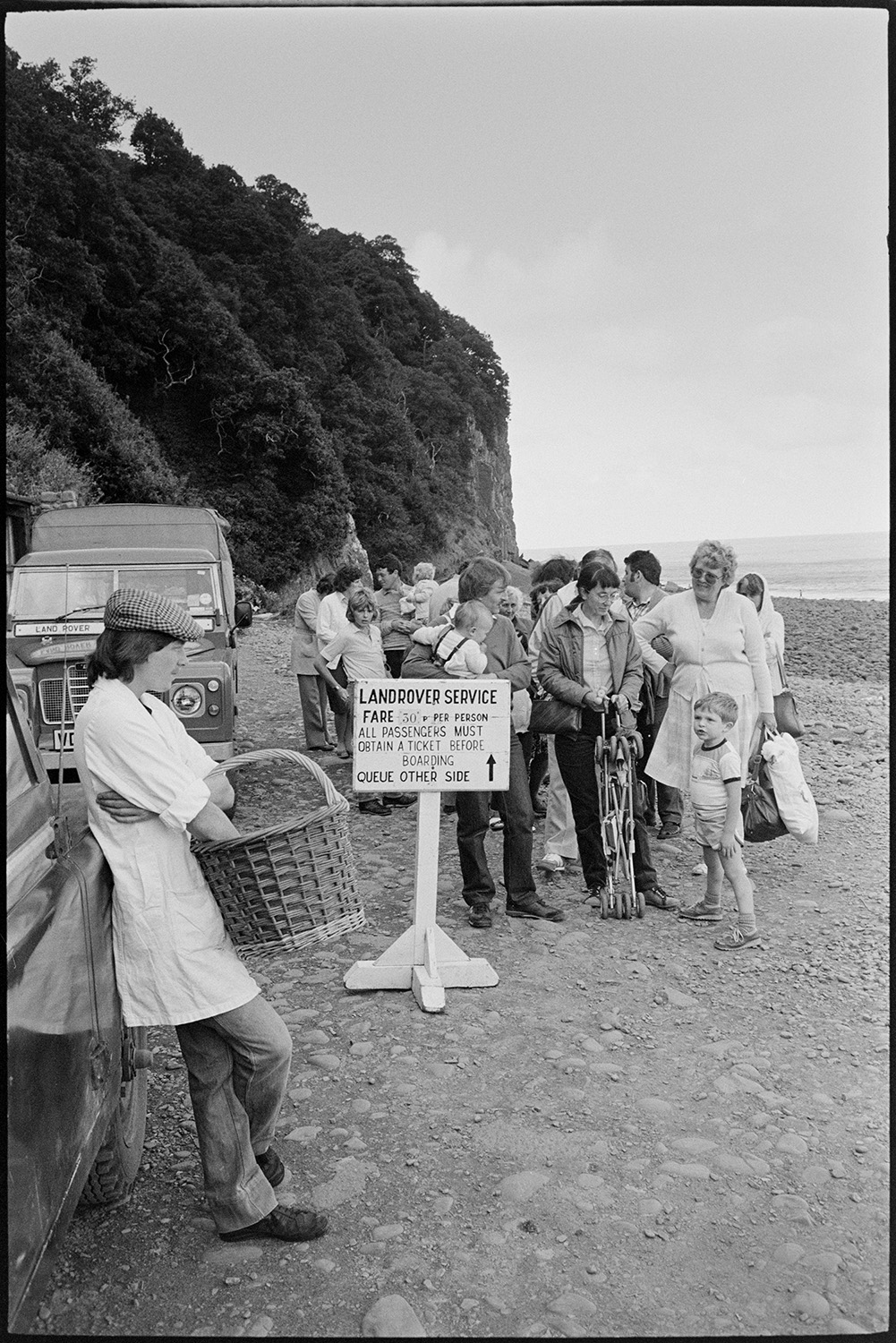 Holidaymakers, seaside village taking photographs, sunbathing, pebble beach with small boats. 
[Holidaymakers queuing up on the pebble beach at Clovelly to get the Land Rover service back through the village. A person holding a wicker basket is leaning against one of the Land Rovers.]