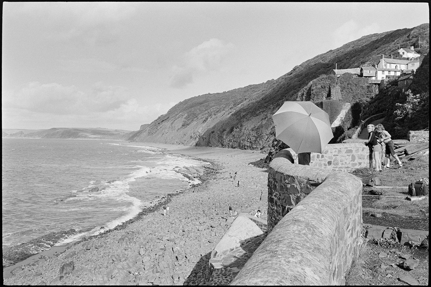 Sea wall with holidaymakers at old harbour, coastal view. 
[Holidaymakers looking over the sea wall to the pebble beach and sea below at Bucks Mills. One person is holding an umbrella and people can be seen walking on the beach below. Houses and the cliffs can be seen in the background.]