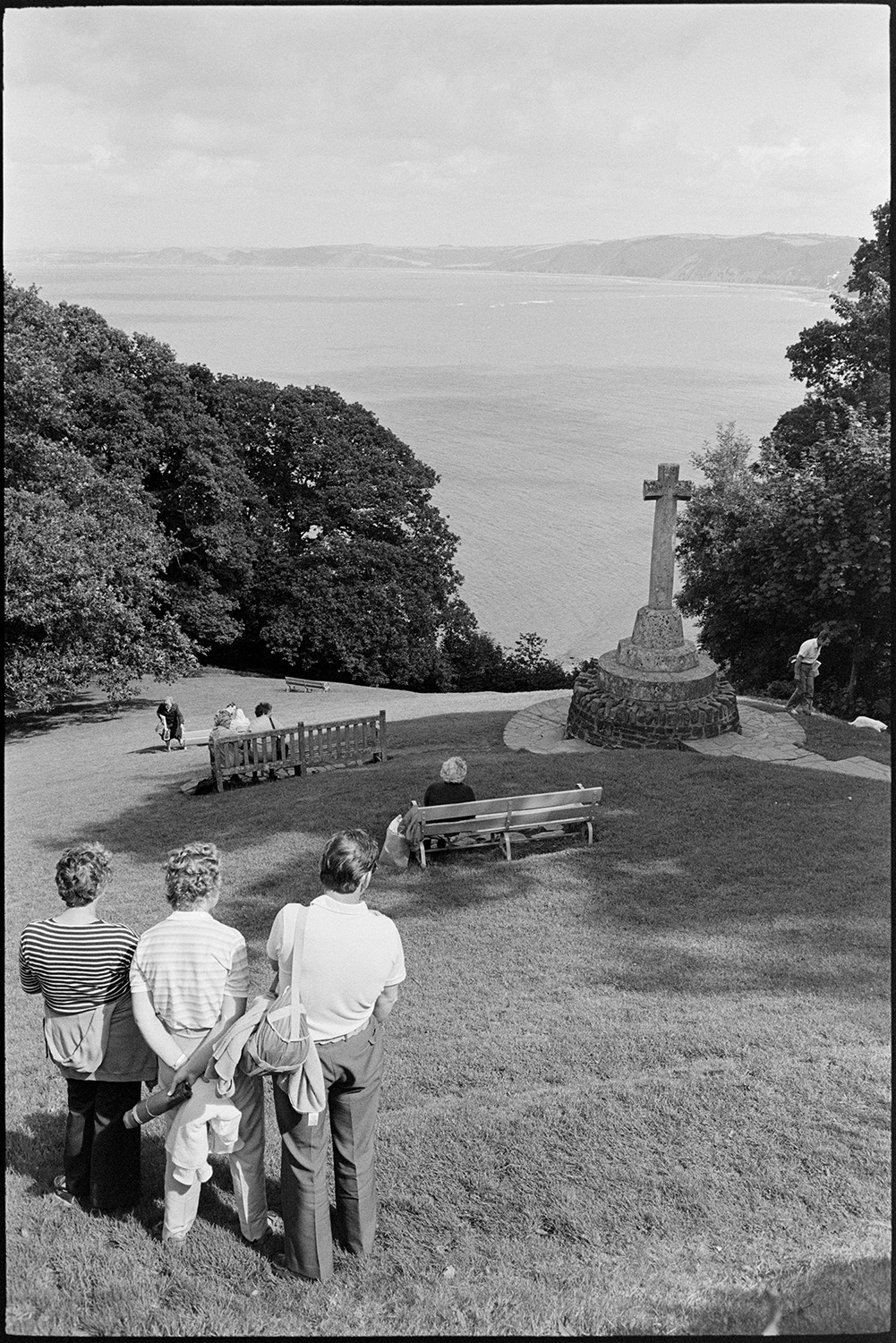 Holidaymakers sitting in park above sea looking at view of bay and coast, stone cross. 
[Holidaymakers sitting on a bench and standing on the grass in a park overlooking the sea and bay at Clovelly. The war memorial for the village can be seen in the park.]