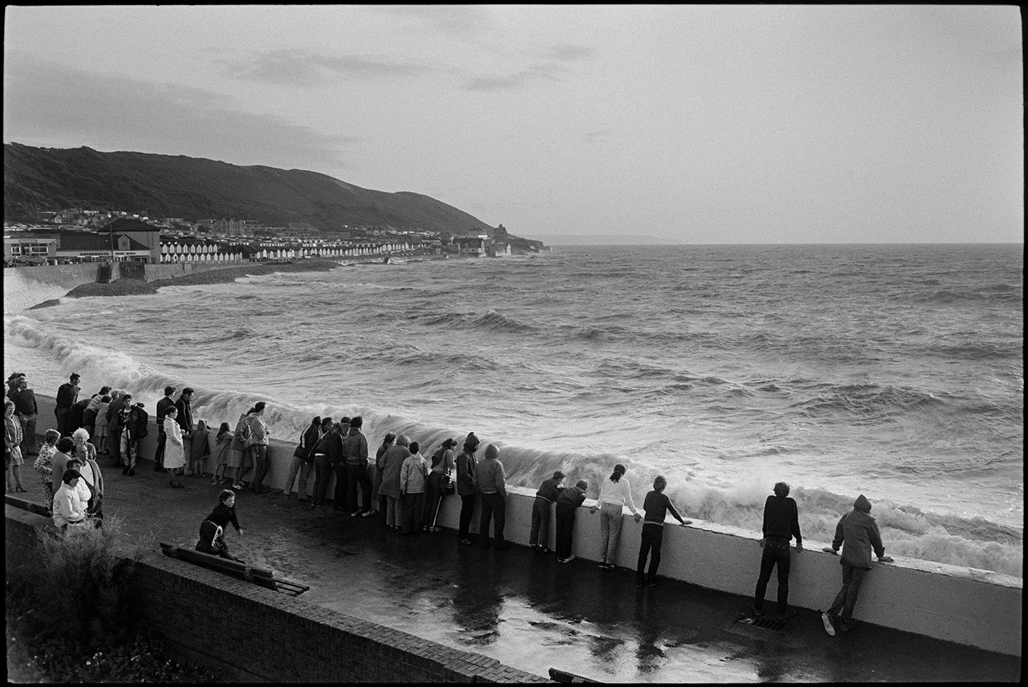 Holidaymakers watching rough sea breaking over sea wall, dodging waves. 
[Holidaymakers watching waves break against the seawall at Westward Ho!. They are lined up along the wall. The town of Westward Ho! can be seen in the background.]
