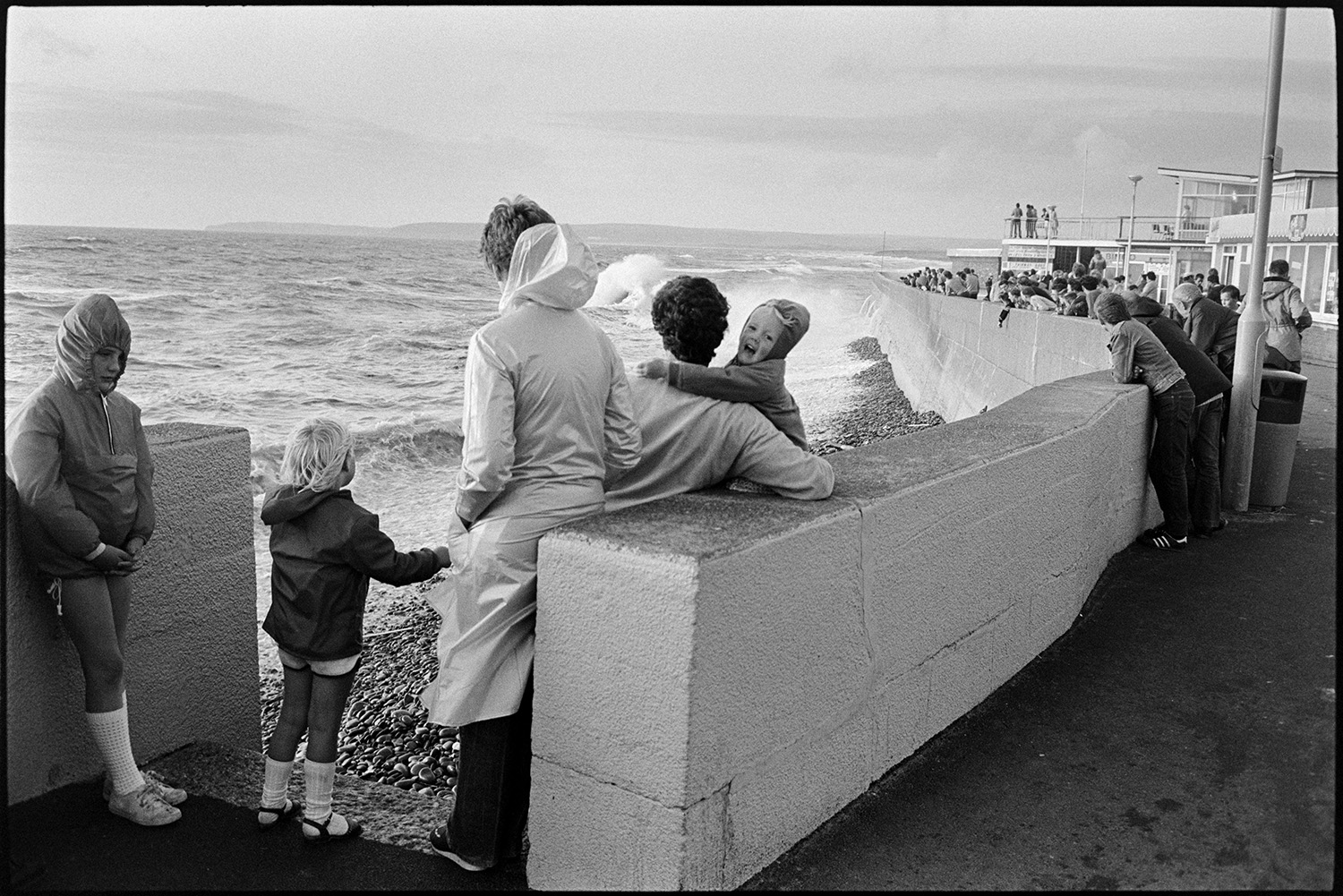 Holidaymakers watching rough sea breaking over sea wall, dodging waves. 
[Holidaymakers watching waves break against the sea wall at Westward Ho!.]