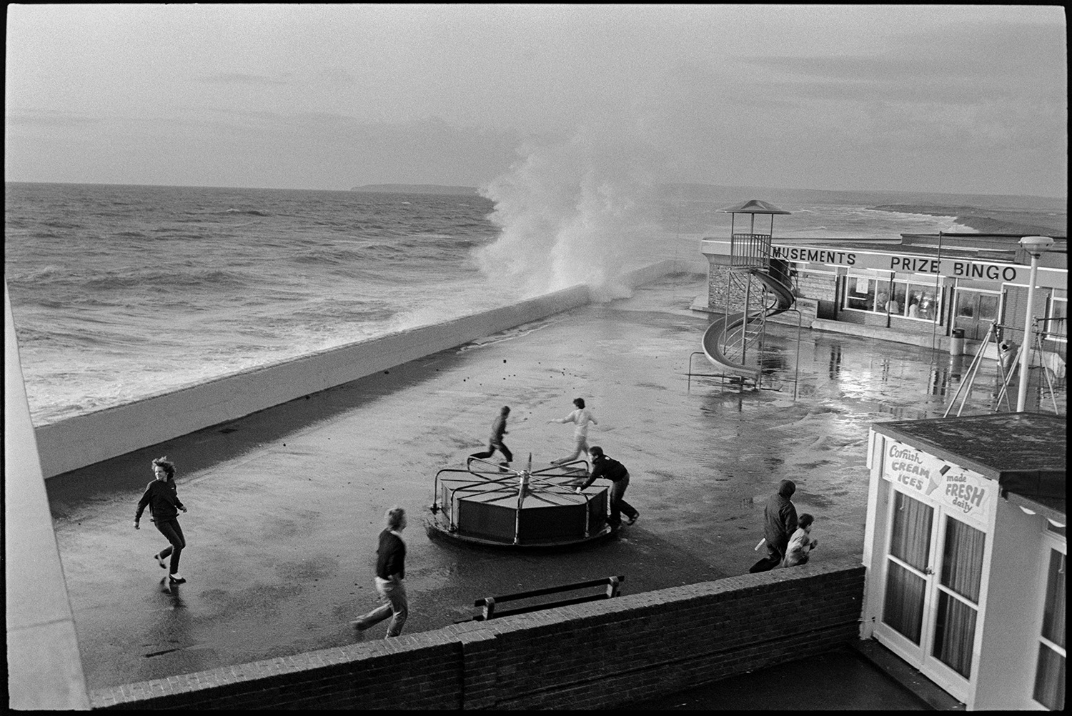 Holidaymakers watching rough sea breaking over sea wall, dodging waves. 
[Teenagers by a roundabout and amusement arcade on the seafront at Westward Ho!. A wave is breaking against the seawall in the background.]