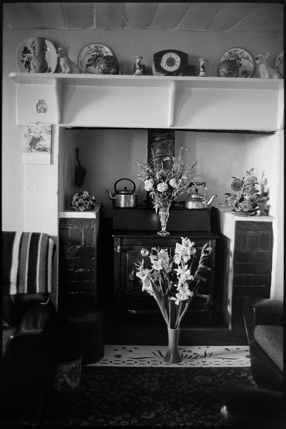 Vases  and displays of flowers by a fireplace in a house at Upcott, Dolton. Two kettles are also displayed on the fire. China plates and ornaments are on display on the mantelpiece above the fireplace.]