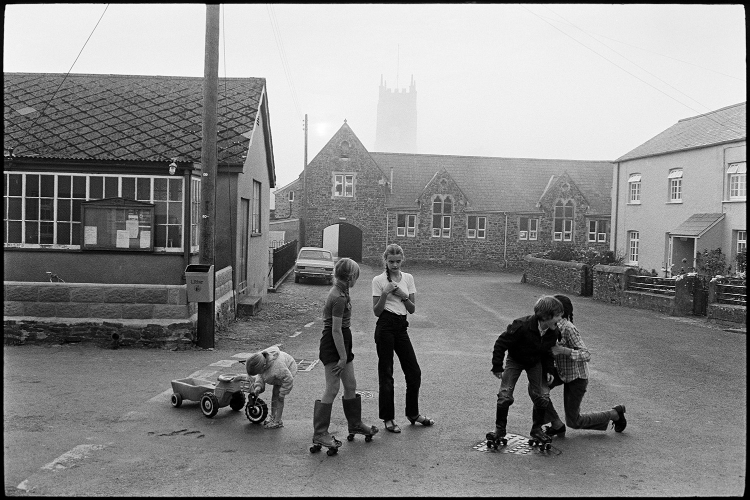Children playing in village street with church tower and Post Office. Misty day, fog.
[Street scene with five children playing in a road at High Bickington. Two children are wearing wellington boots with roller skates and a young child has a ride on toy tractor and trailer. A building with a notice board is on the street corner and the church tower can be seen in the mist and fog in the background.]