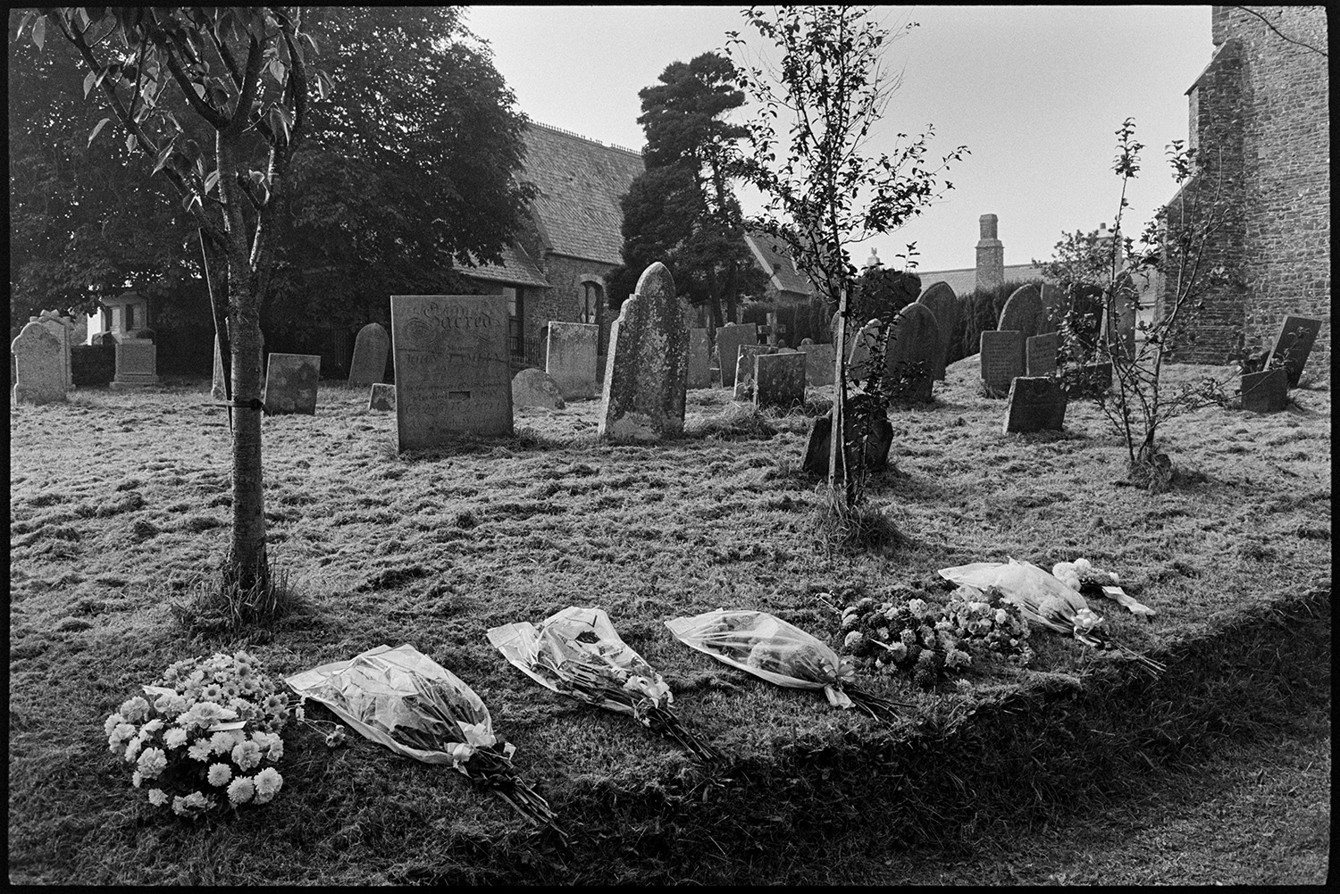 Bunches of funeral flowers in graveyard after funeral.
[Arrangements and bouquets of flowers laid out along the grass bank by gravestones in Atherington churchyard, after a funeral.]