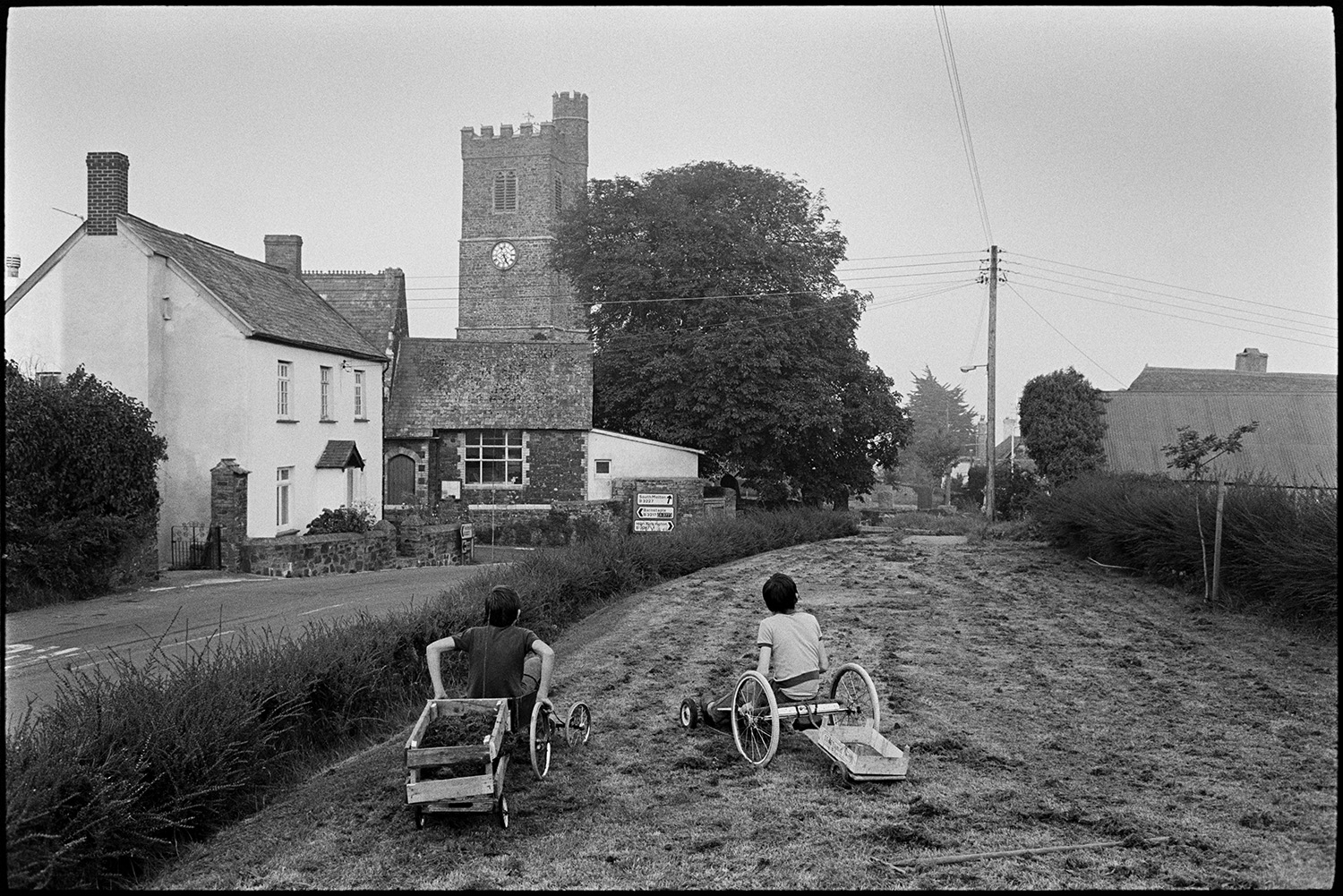 Children playing with buggy and trying to knock down conkers from chestnut tree, church.
[Two children sitting on home made go-karts in a small field of newly cut grass by the road opposite the church in Atherington.]