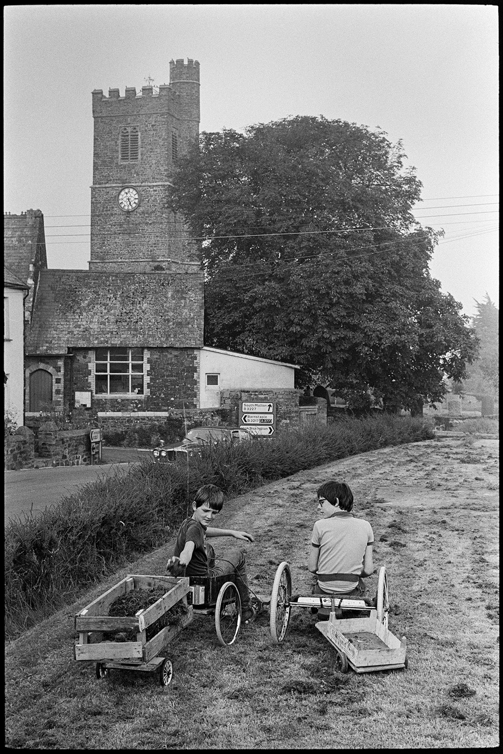 Children playing with buggy and trying to knock down conkers from chestnut tree, church.
[Two boys sitting on home made go-karts in a small field of newly cut grass by the road opposite the church in Atherington. One boy is collecting the grass cuttings in his trailer.]