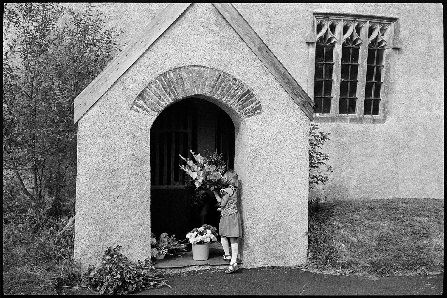 Women decorating church for Harvest Festival, flowers.
[A girl in the porch at Dowland Church carrying a big bouquet of flowers for the Harvest Festival decorations. A bucket with flowers and a pile of foliage are at the church entrance.]