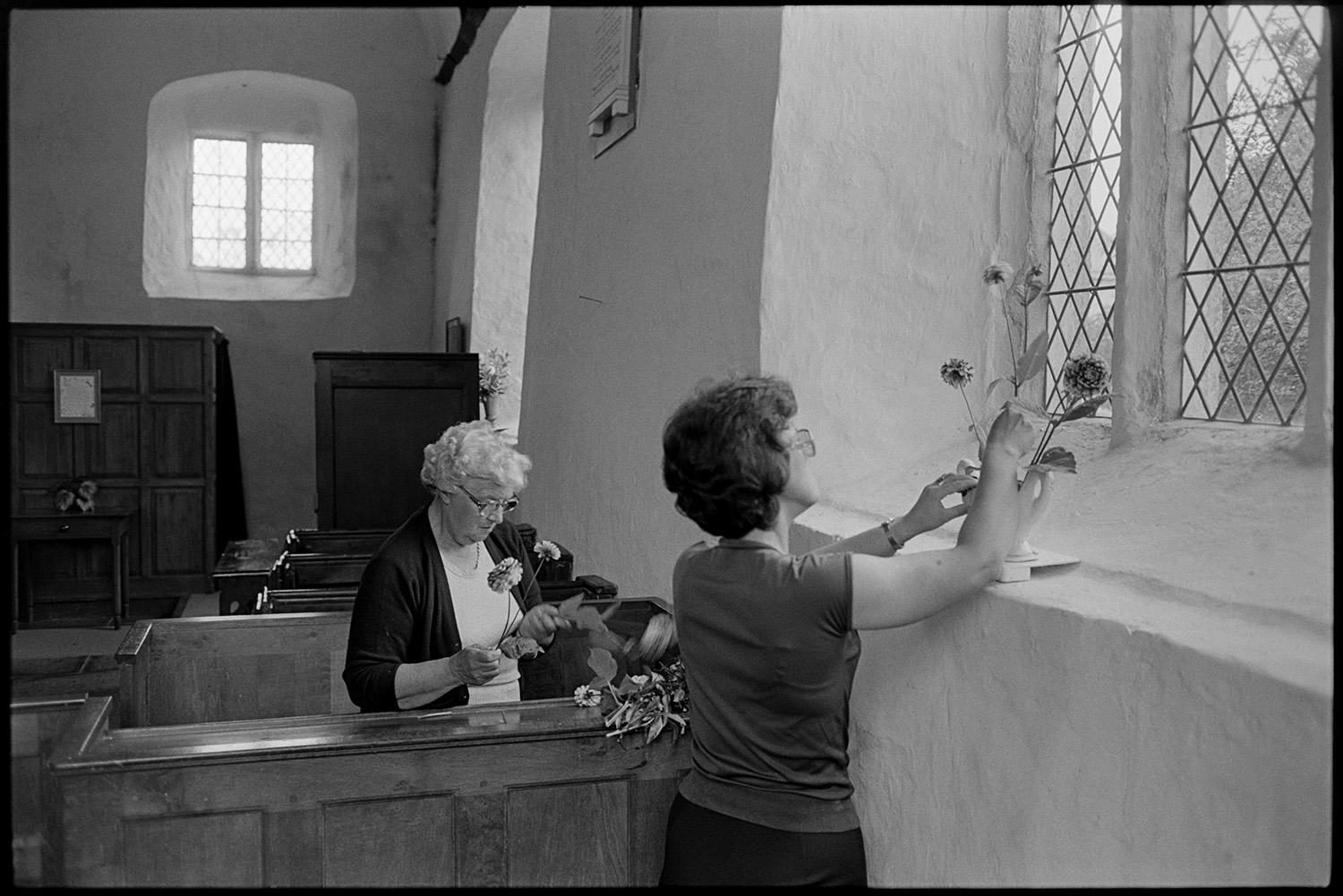Women decorating church for Harvest Festival. Flowers.
[Two women arranging flowers in a vase on a windowsill in Dowland Church for the Harvest Festival. The women are stood in wooden pews.]