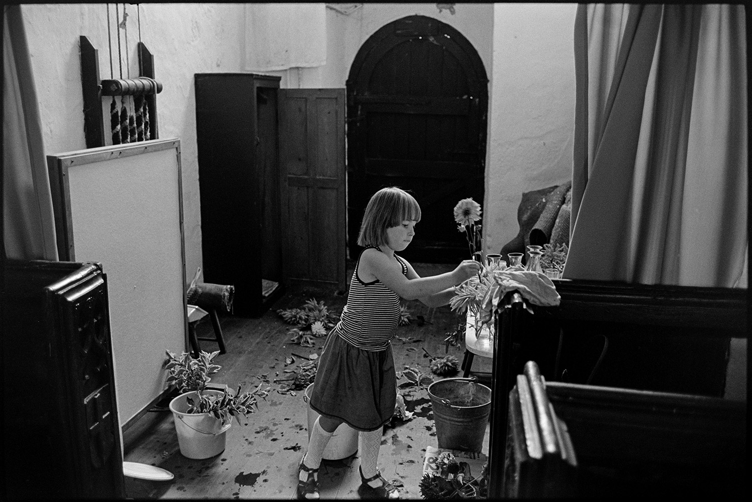Women decorating church for Harvest Festival. Flowers.
[A girl surrounded by buckets and cut flowers in Dowland Church, arranging flowers in a jar for the Harvest Festival. Wooden pew ends can be seen in the foreground and bell ropes are visible against a wall behind her.]