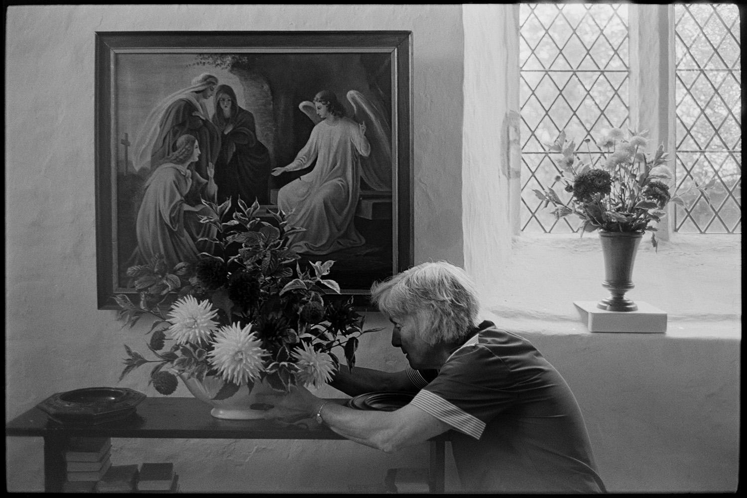 Women decorating church for Harvest Festival, flowers. Religious painting on wall. 
[Evelyn Folland making a floral decoration in front of a religious painting on the wall for the Harvest Festival at Dowland Church.]