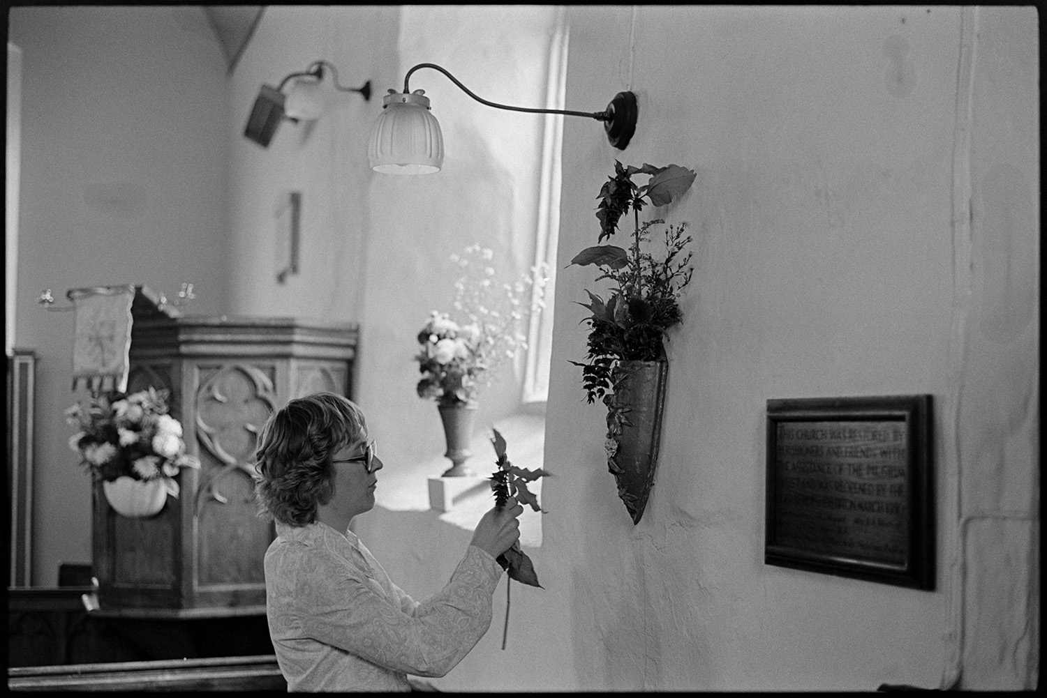 Women decorating church for Harvest Festival, flowers. Religious painting on wall. 
[Mrs Squire making a floral decoration on a wall by a plaque, at Dowland Church for the Harvest Festival. The pulpit and other flower arrangements can be seen in the background.]