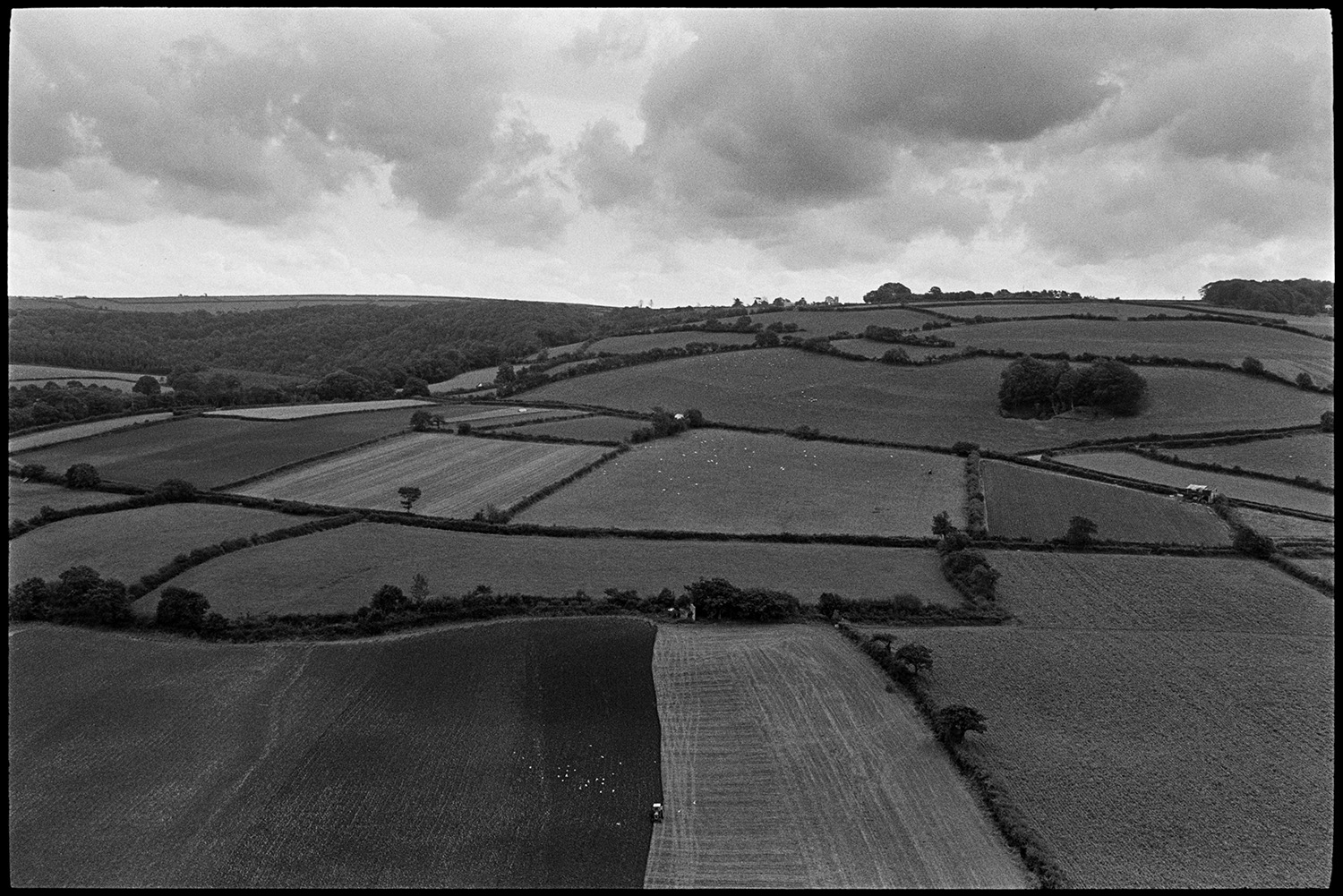 Landscape with ploughed fields clouds and hedgerows.
[A view of hills with woodland, fields and hedges at Torrington. A tractor can be seen ploughing a field in the foreground. Clouds are visible in the sky above the fields.]
