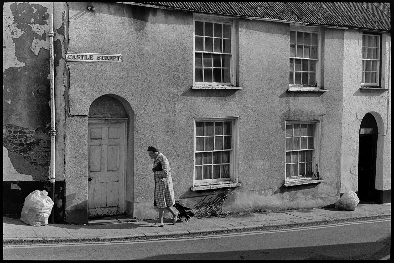 Town street with fine house front, passers by on pavement nice windows, round arched door.
[A woman, pulling a shopping trolley, walking past a house at Castle Street, Torrington. A bag of rubbish is on the pavement by a wall with crumbling plasterwork.]