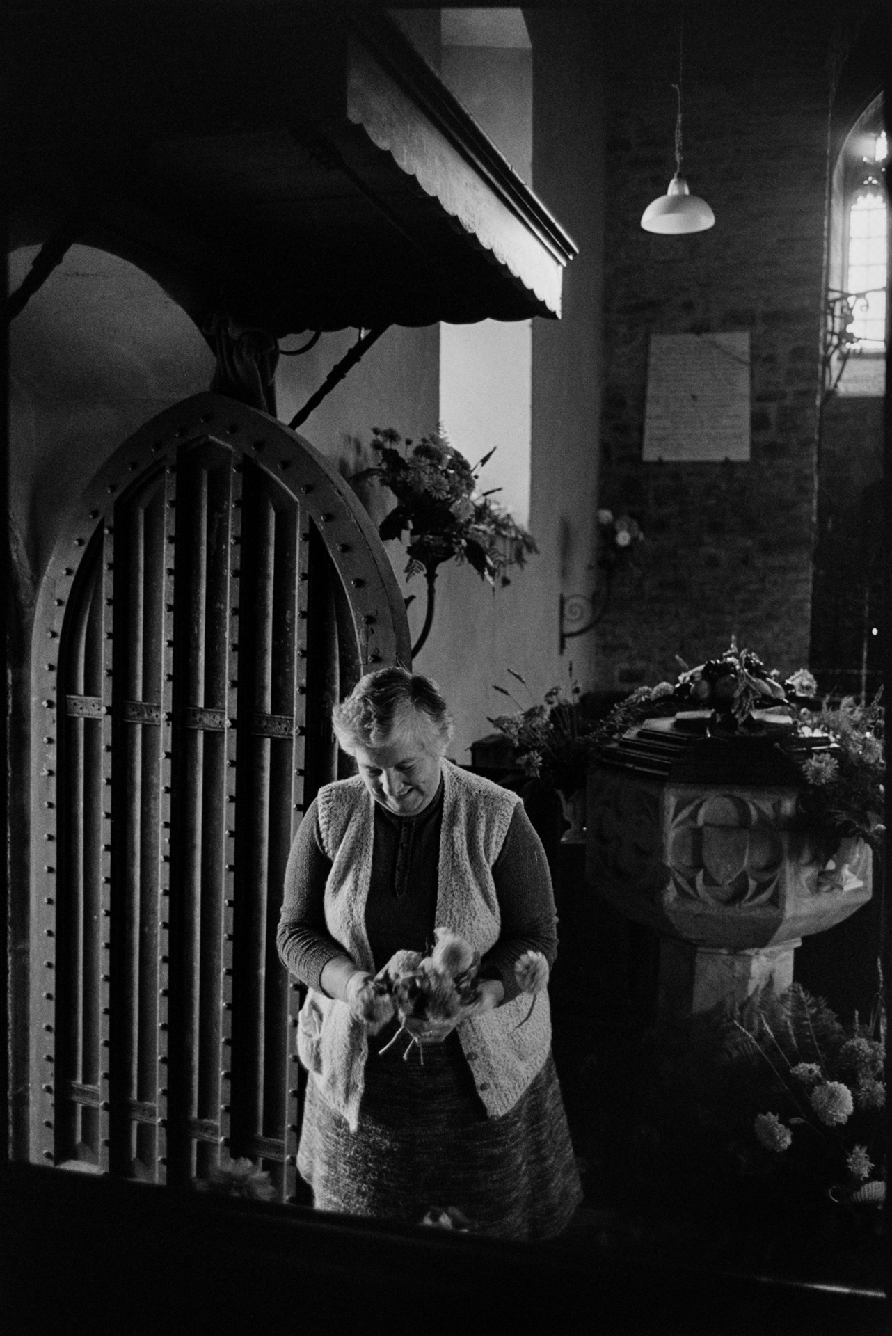 Women decorating church for Harvest Festival, flowers and produce. 
[A woman stood inside the doorway of Atherington Church. She is holding a bunch of flowers to decorate the church for the Harvest Festival. Flower displays can be seen on the font behind her.]