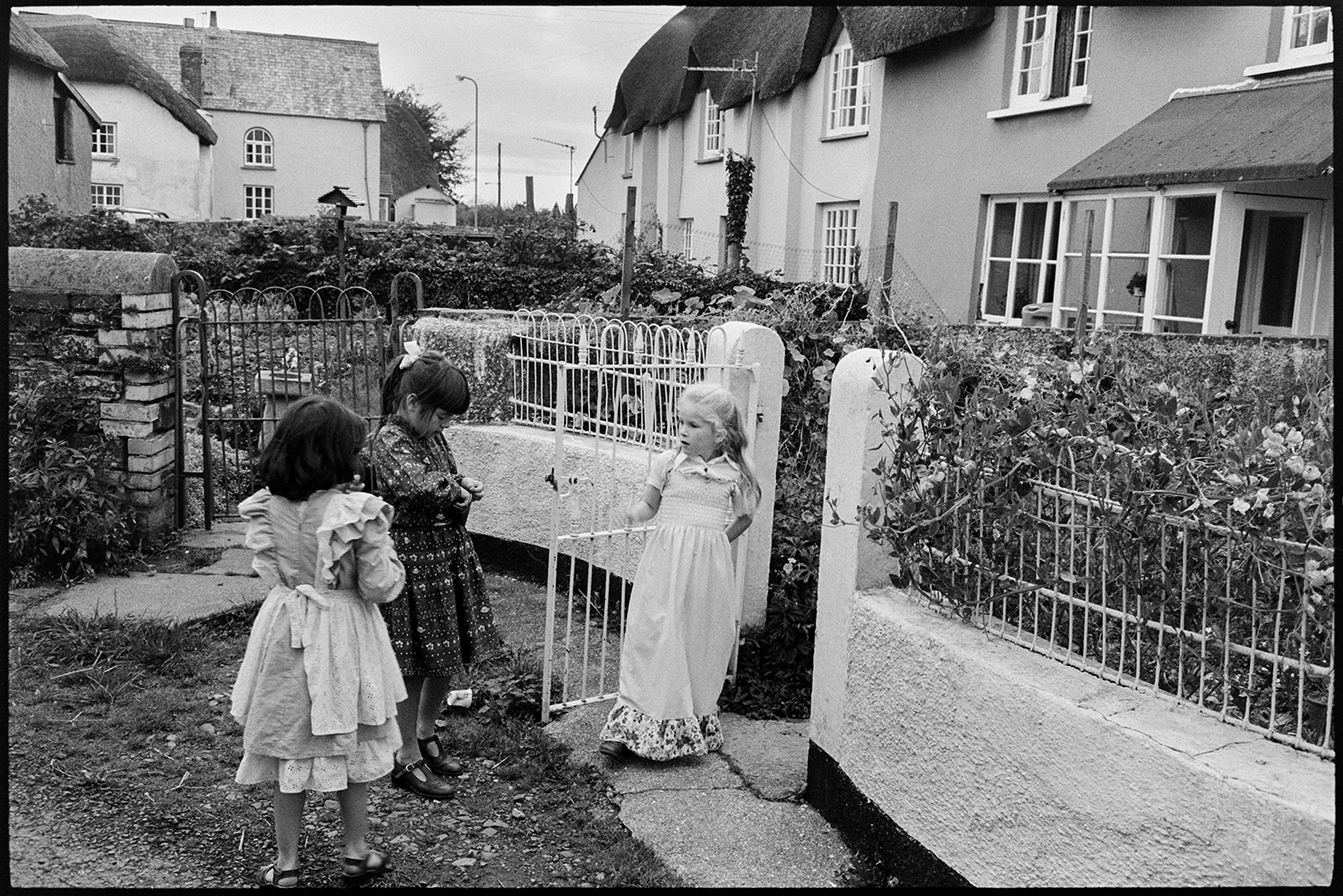 Birthday party with children in party dresses. 
[Three girls in party dresses by railings outside the thatched cottage of Quillets, Dolton, for Hannah Bignell's birthday party. The cottage was previously known as Park View.]