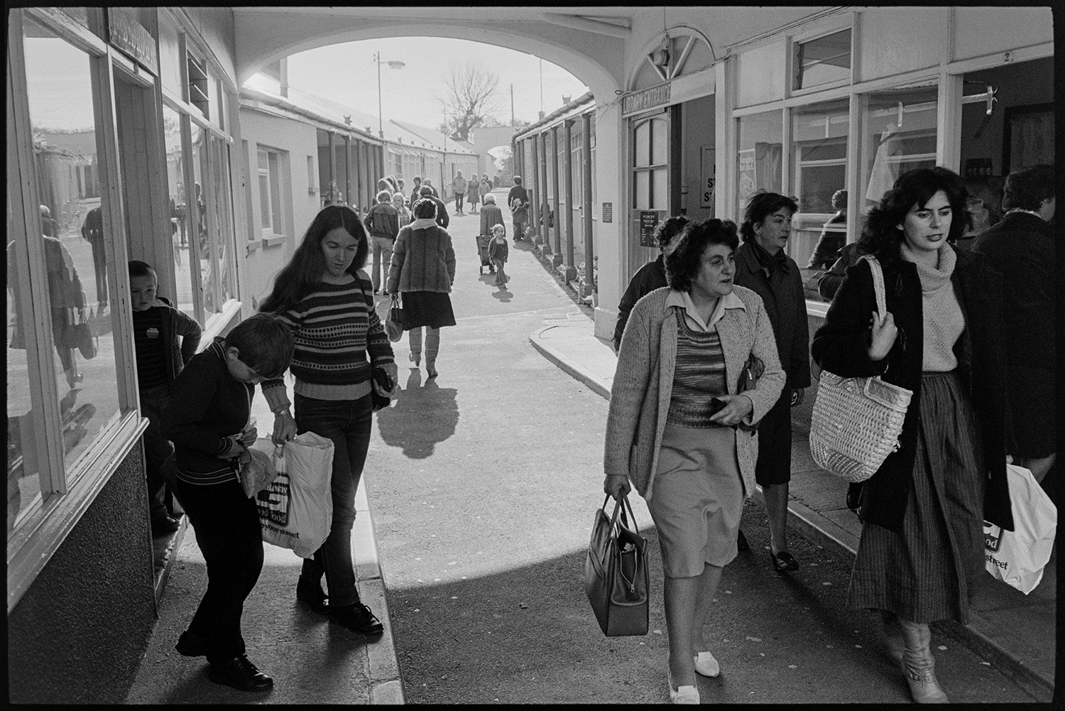 Shoppers in market row. 
[Shoppers walking along Market Row, Torrington. A woman and two children are coming out of a shop doorway.]