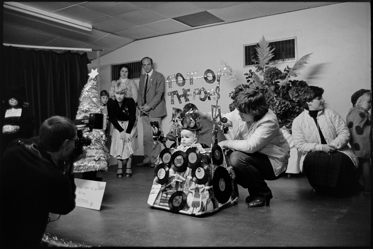 Village carnival, children's fancy dress competition in village hall. 
[A man taking photographs of entrants in a children's fancy dress competition for Dolton Carnival in Dolton Village Hall. He is photographing a toddler dressed as 'Tot of the Pops'.]
