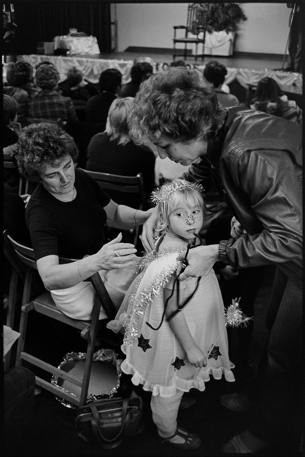 Village carnival, children's fancy dress competition in village hall. 
[Two women adjusting the wings of a child dressed as a fairy at a fancy dress competition in Dolton Village Hall for Dolton Carnival.]