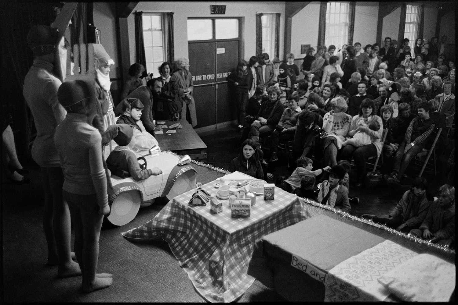 Fancy dress competition in village hall, children's King and Queen on stage. 
[A crowd of people in Dolton Village Hall watching a children's fancy dress parade for Dolton Carnival. One child is dressed as Noddy. A model bed and table laid with breakfast is also on the stage.]