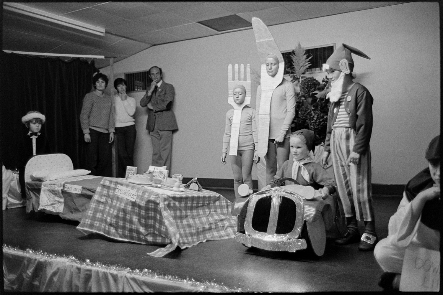Fancy dress competition in village hall, children's King and Queen on stage. 
[Children in a fancy dress competition at Dolton Village Hall for Dolton Carnival. Two children are dressed as Noddy and Big Ears, while two other children are dressed as a knife and fork next to a model bed and table laid for breakfast.]