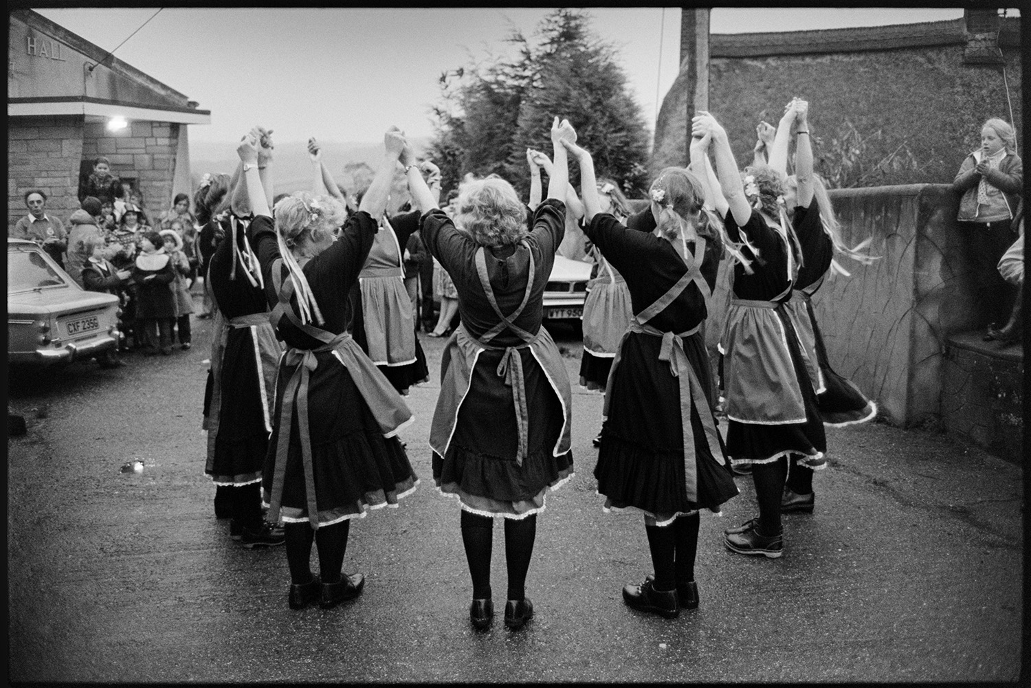 Women's Clog Dancing team at village carnival. 
[A group of women clog dancers performing in a circle outside Dolton Village Hall at Dolton Carnival. People are watching them.]
